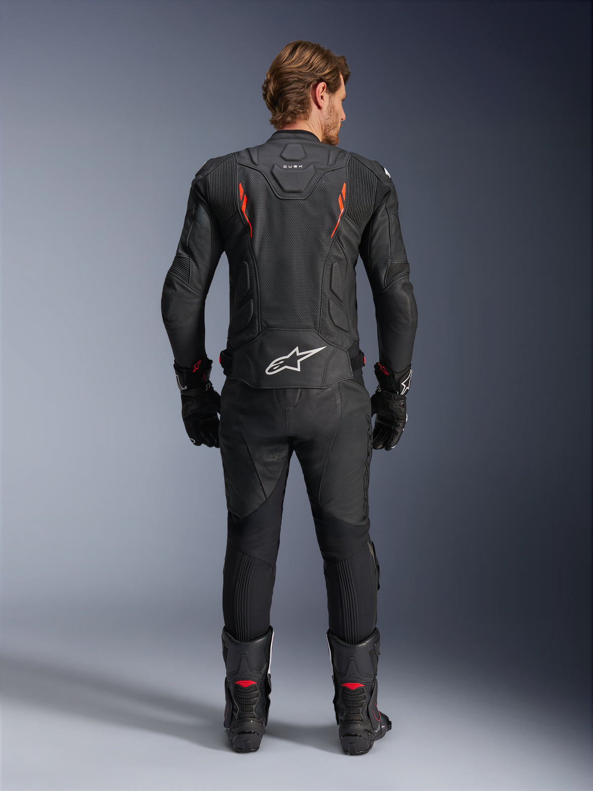 Alpinestars Dusk Airflow Leren Jas, zwart en fluorood, achteraanzicht met uitgebreide perforaties en groot wit logo op de onderrug, gedragen met bijpassende leren race-uitrusting inclusief broek, laarzen en handschoenen