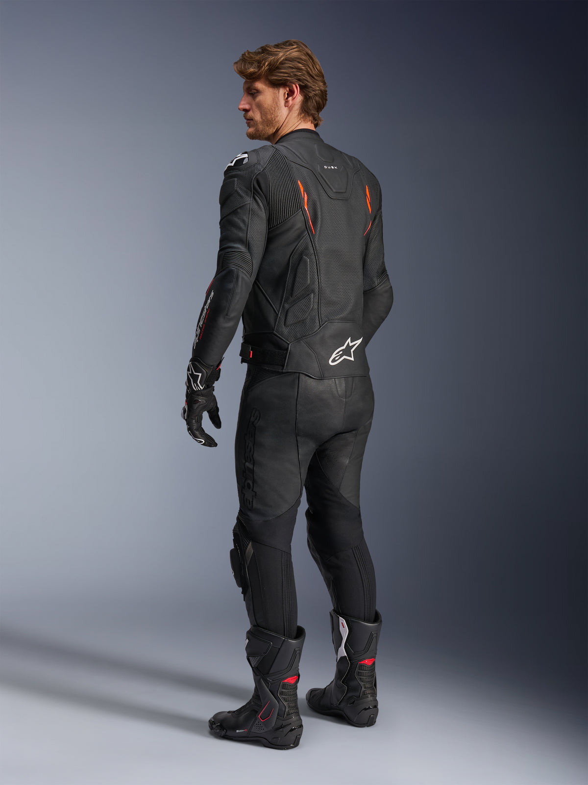 Alpinestars Dusk Airflow Leren Jas, zwart met Red Fluo accenten, achteraanzicht van een rijder in volledige leren motoruitrusting inclusief bijpassende zwarte broek, handschoenen en racelaarzen, voorzien van geperforeerd leer en witte Alpinestars-logo's.