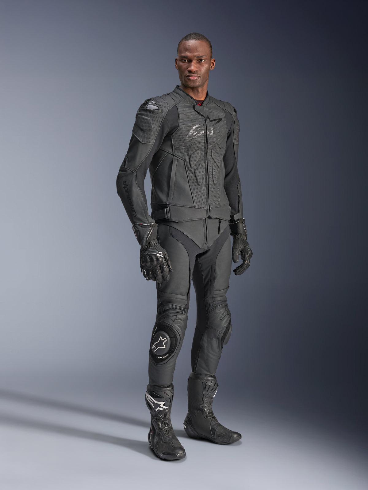 Veste en cuir Alpinestars Dusk Airflow, noir, portée par un pilote avec un équipement technique complet comprenant un pantalon en cuir assorti, des gants et des bottes, avec des panneaux en cuir perforé pour la ventilation et une armure intégrée pour la conduite moto sportive