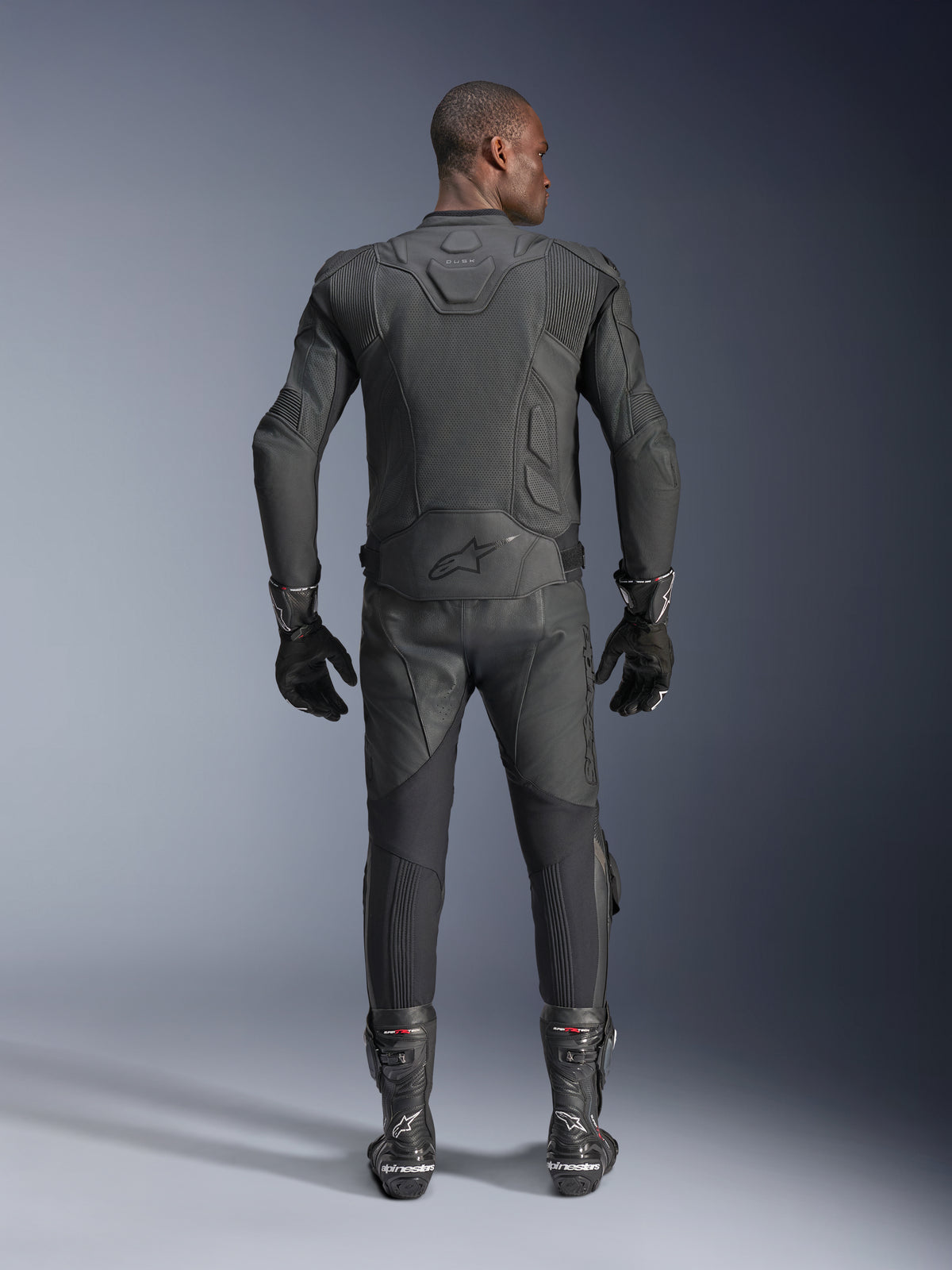 Modèle masculin vu de dos portant la veste en cuir Alpinestars Dusk Airflow noire, associée à un pantalon de course, des gants et des bottes en cuir noir assortis, avec des perforations étendues pour la ventilation et le logo Alpinestars dans le bas du dos.