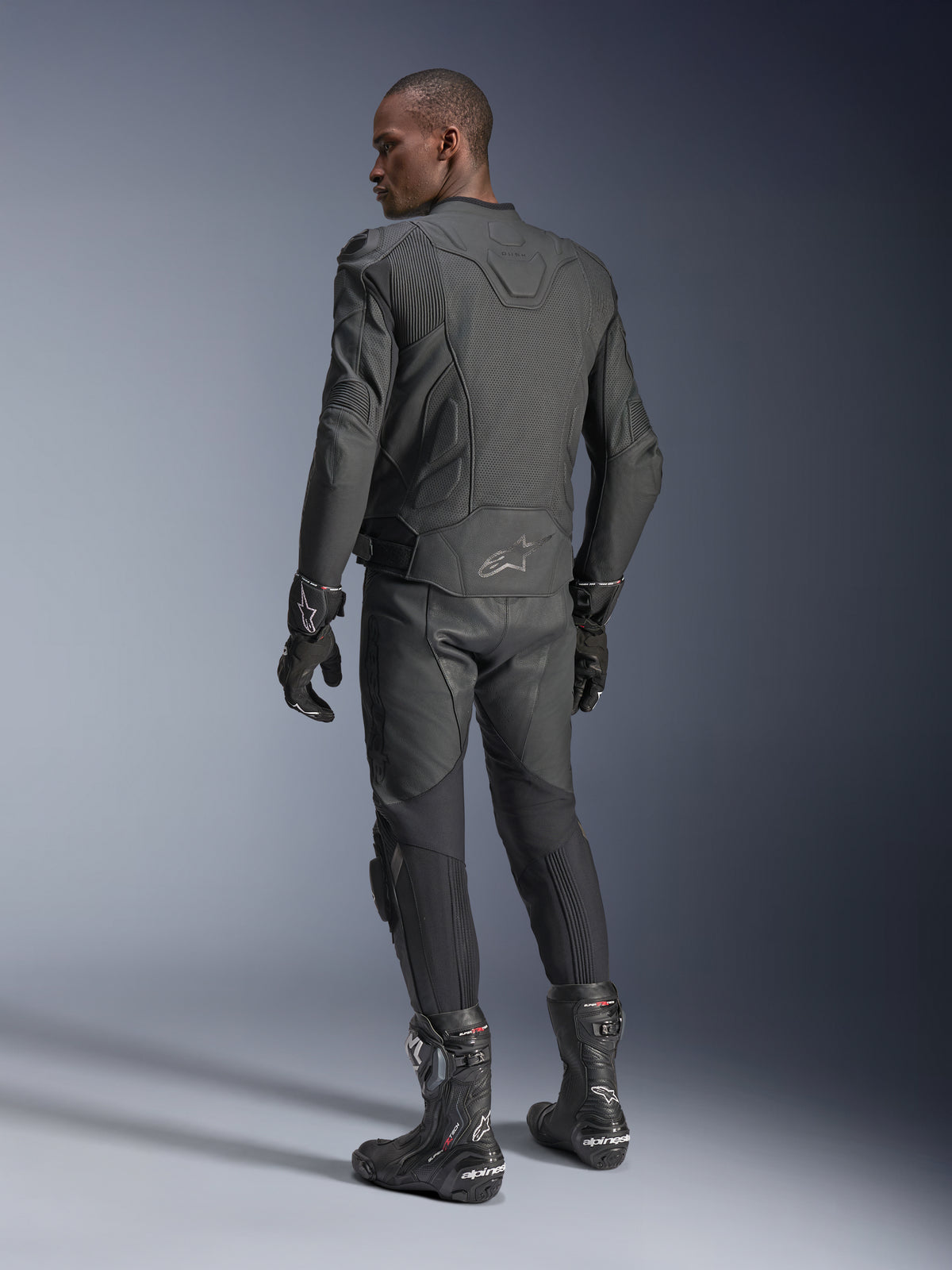 Pilote de dos portant la veste en cuir Alpinestars Dusk Airflow en Black Black, avec pantalon en cuir, gants et bottes assortis, mettant en avant les perforations étendues au dos et la coupe sport haute performance.