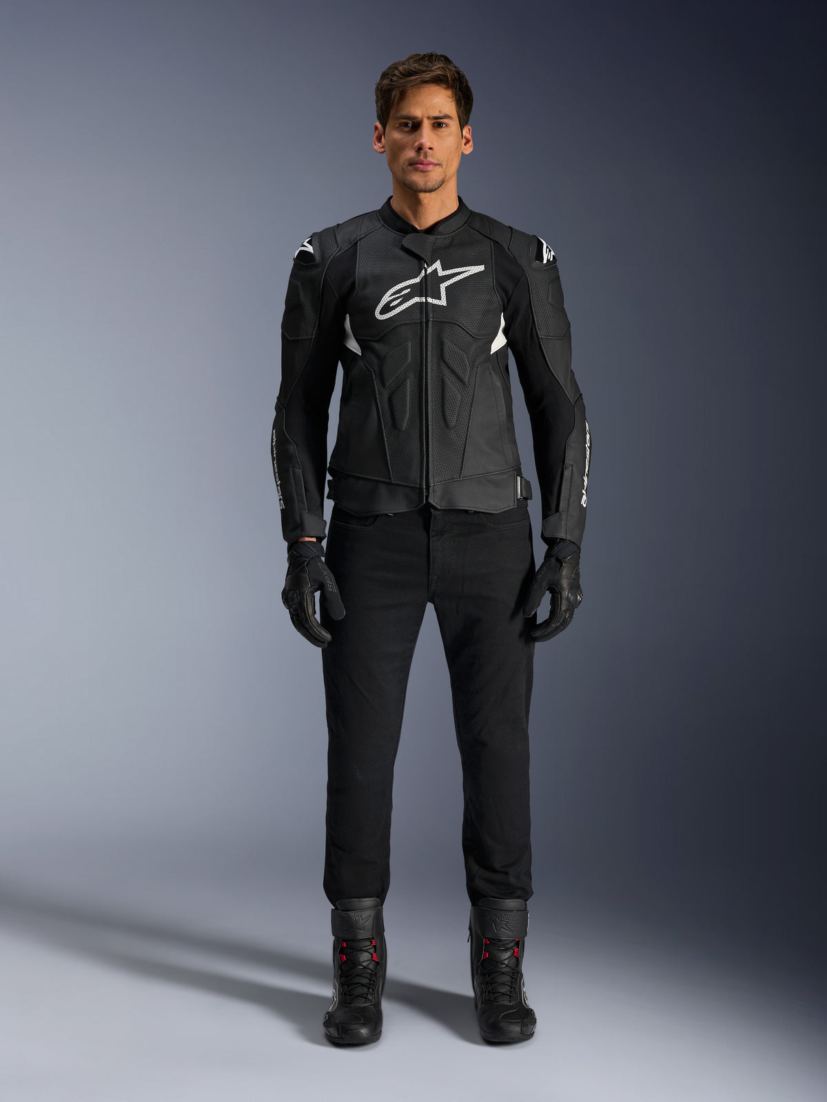 Veste en cuir Alpinestars Dusk Airflow, noir et blanc, veste de moto en cuir avec panneaux perforés pour la circulation de l'air, logo Astars blanc sur la poitrine, portée par un pilote avec pantalon noir, gants et bottes de moto, photo professionnelle en studio pour équipement de moto de sport.