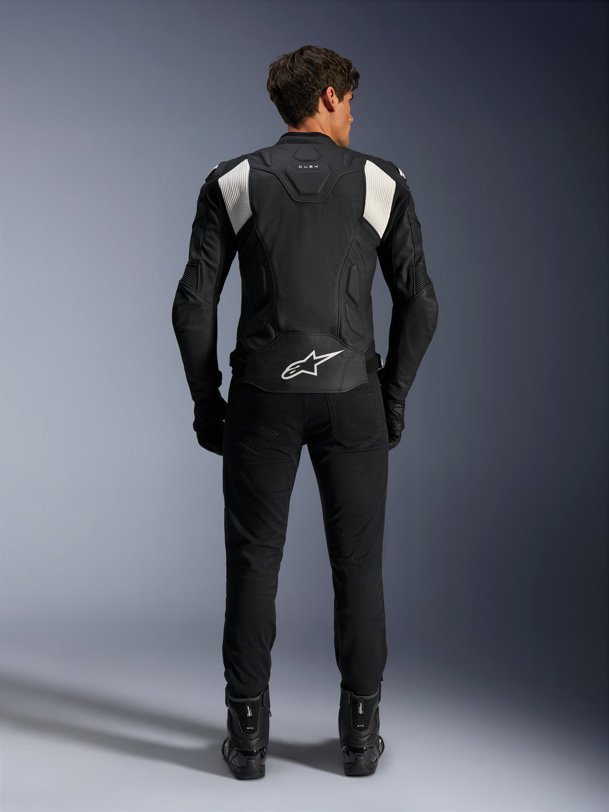 Veste en cuir Alpinestars Dusk Airflow, noir et blanc, vue arrière d'un pilote portant un pantalon et des bottes noirs, dotée de cuir perforé pour la ventilation, de panneaux stretch en accordéon blancs sur les épaules et d'un logo blanc sur le bas du dos
