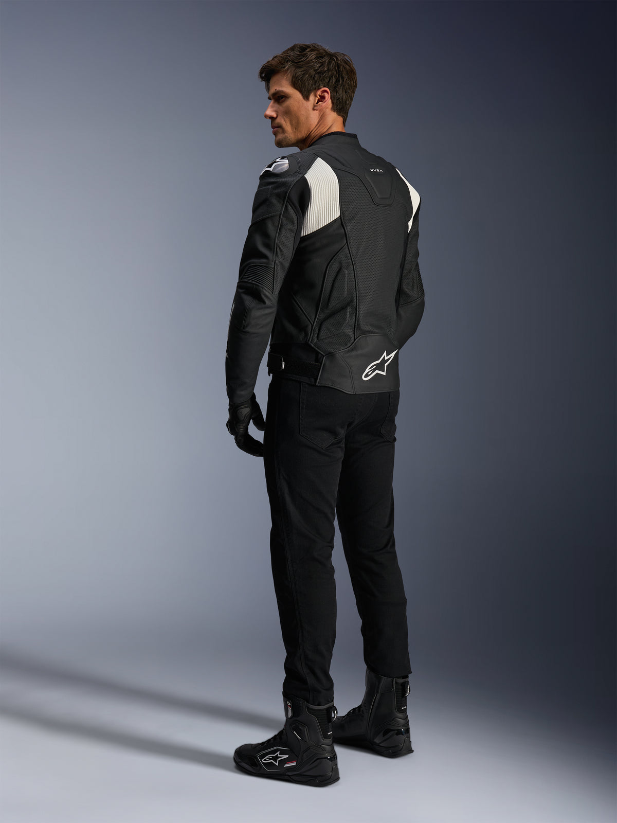 Veste en cuir Alpinestars Dusk Airflow, noir et blanc, vue arrière d'un mannequin portant la veste en cuir perforé avec pantalon et bottes noirs assortis, présentant des panneaux extensibles en accordéon blancs et un logo imposant dans le bas du dos, équipement de moto sport