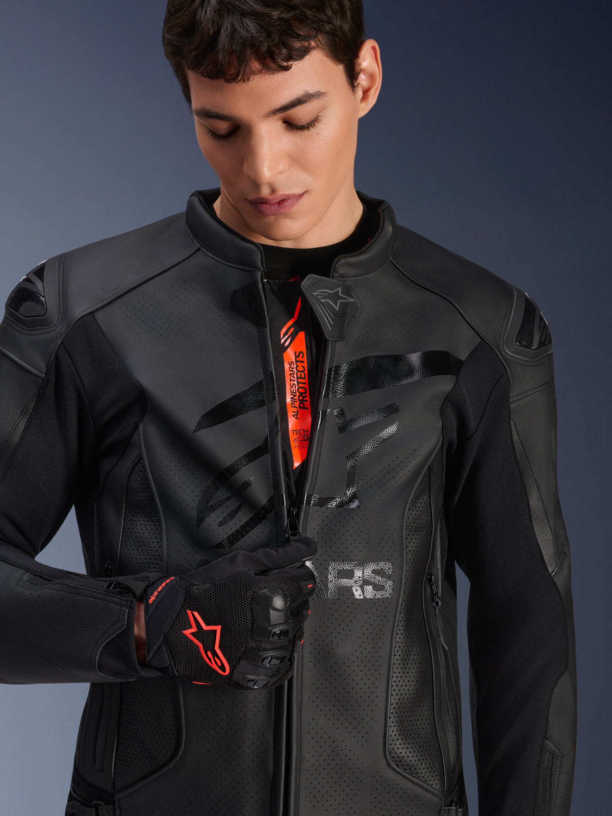 Alpinestars GP Plus R V4 Airflow Leather Jacket in Black, male model partially unzipping the front to show orange internal branding, featuring perforated leather panels and a glossy chest logo, paired with a black and red motorcycle glove
