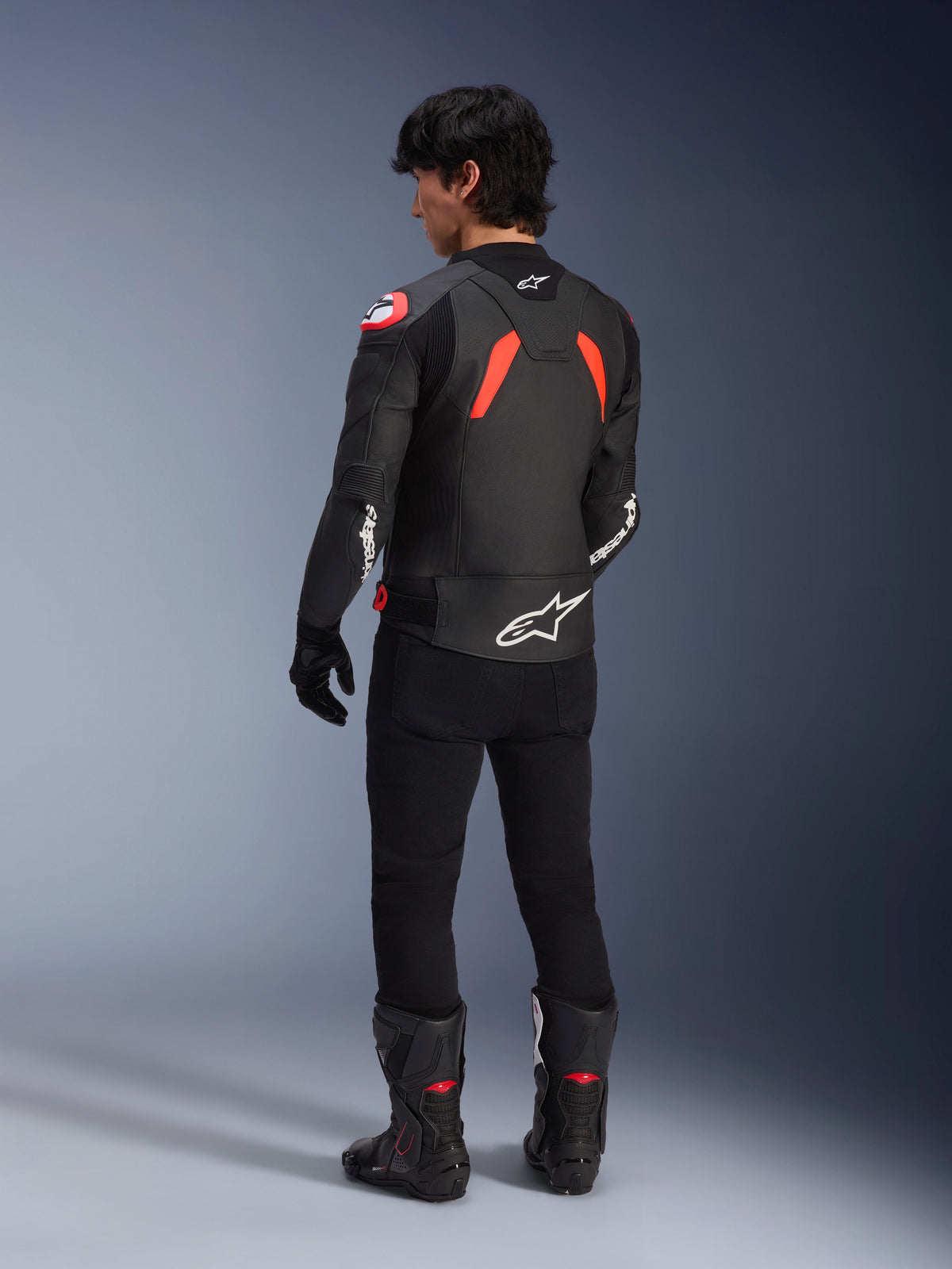 Vue arrière d'un mannequin portant la veste en cuir Alpinestars GP Plus R V4 Airflow, noire avec des accents rouge fluo et blancs, portée avec un pantalon noir et des bottes de moto noires, logos blancs proéminents sur le dos et les manches, construction en cuir de style sportif