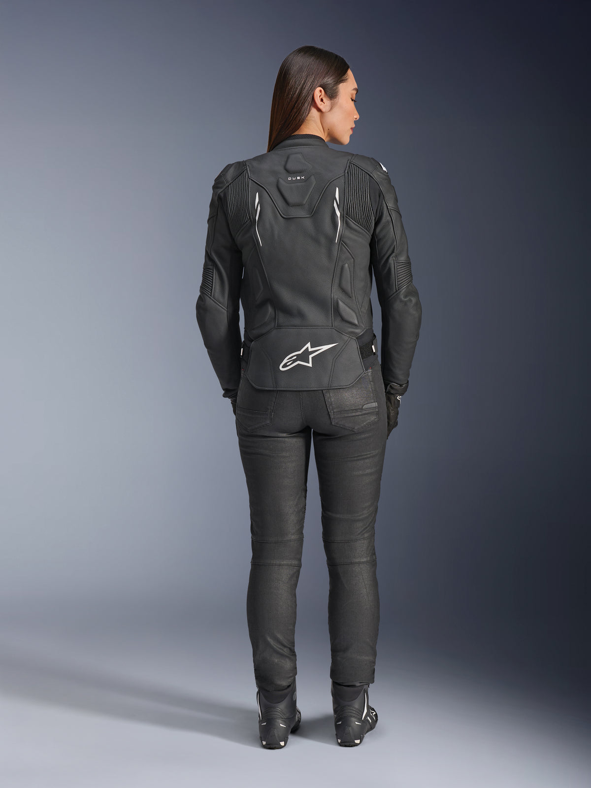 Alpinestars Stella Dusk leren jas voor dames, zwart, achteraanzicht met wit Alpinestars-logo op de onderrug en 'DUSK'-branding op de bovenrug, gedragen met zwarte motorbroek en laarzen, toont de aansluitende damespasvorm en beschermende leren constructie.