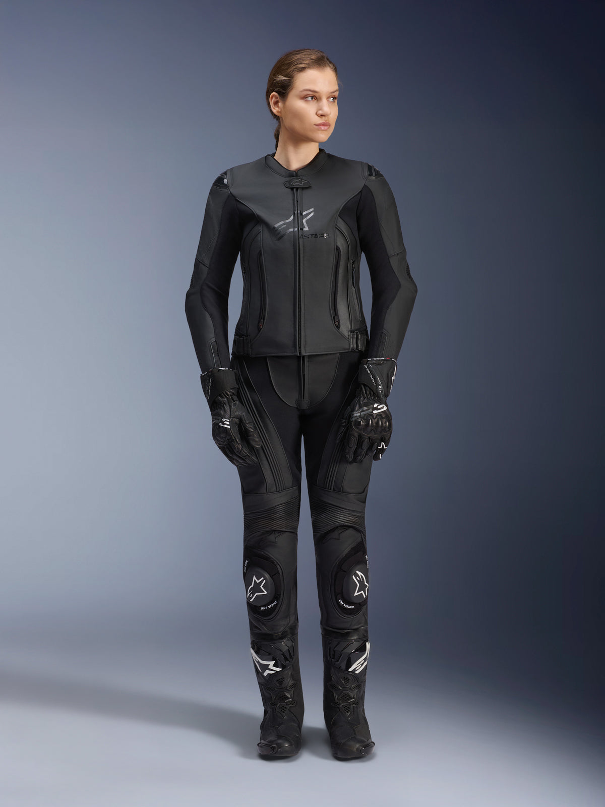Alpinestars Damen Stella GP Plus V4 Lederjacke in Schwarz, getragen von einer Motorradfahrerin mit passender Leder-Rennhose, Handschuhen und Stiefeln, taillierte Passform, zentraler Reißverschluss und dezentes Logo auf der Brust, koordiniert für Sport- und Straßenmotorradfahren.