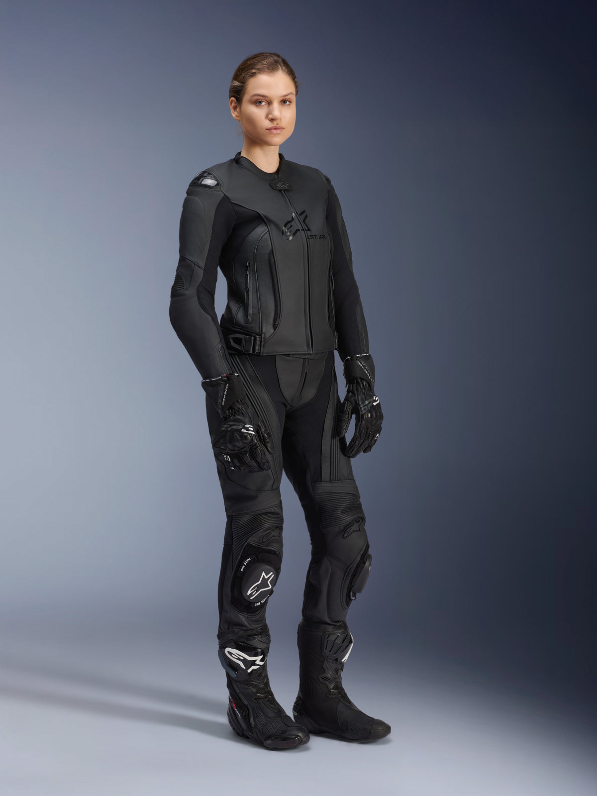 Motorradfahrerin in der Alpinestars Stella GP Plus V4 Damen-Lederjacke in Schwarz Schwarz, gestylt als kompletter Leder-Rennanzug mit passender Motorradhose, Handschuhen und Stiefeln, elegantes Performance-Design mit integriertem Schutz