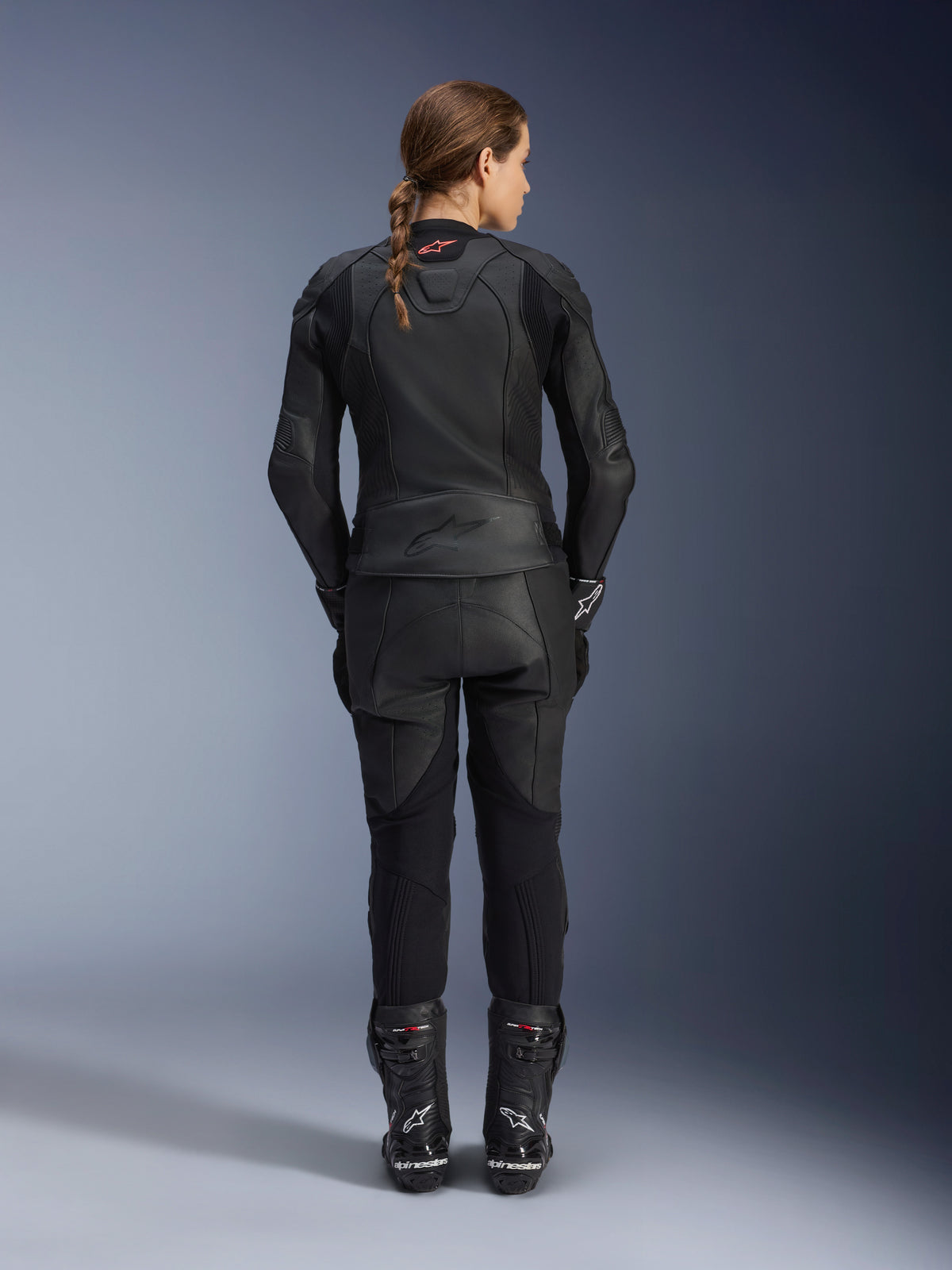 Alpinestars Stella GP Plus V4 Damen-Lederjacke, Schwarz Schwarz, Ganzkörper-Rückansicht einer Fahrerin in passender Lederhose und Stiefeln, elegantes minimalistisches Design mit kleinem roten Logo im Nacken, taillierte Sport-Silhouette für Damen