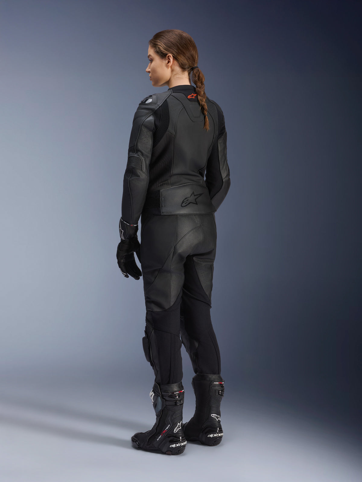 Stella GP Plus V4 Lederjacke für Damen, Schwarz, vollständige Rückansicht, kombiniert mit schwarzer Motorrad-Lederhose, Rennhandschuhen und Stiefeln, mit geprägtem Alpinestars-Logo am unteren Rücken und stromlinienförmiger Silhouette für sportliches Fahren