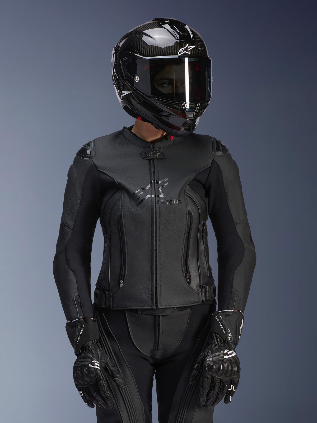 Alpinestars Damen Stella GP Plus V4 Lederjacke in Schwarz, getragen mit schwarzem Carbon-Helm und Rennhandschuhen, elegante Lederkonstruktion mit dezentem Branding auf der Brust, taillierte Silhouette für Fahrerinnen, professionelle Sport-Motorradausrüstung