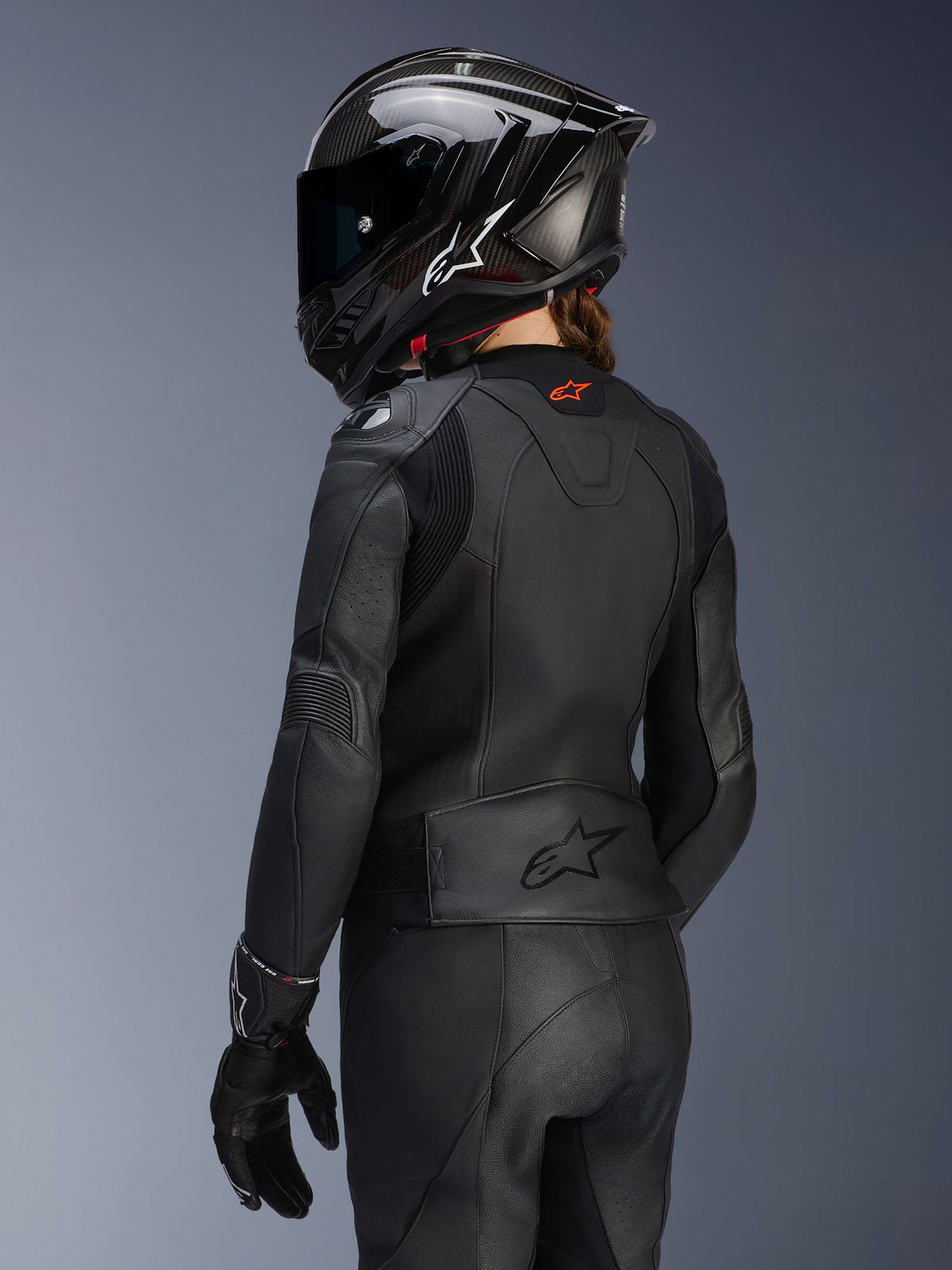 Rückansicht einer Fahrerin in der Alpinestars Stella GP Plus V4 Damen-Lederjacke in Schwarz, mit passender Lederhose, Handschuhen und einem schwarzen Integralhelm im Carbon-Stil; hervorgehoben werden die aerodynamische Passform der Jacke und das geprägte Logo am unteren Rücken.