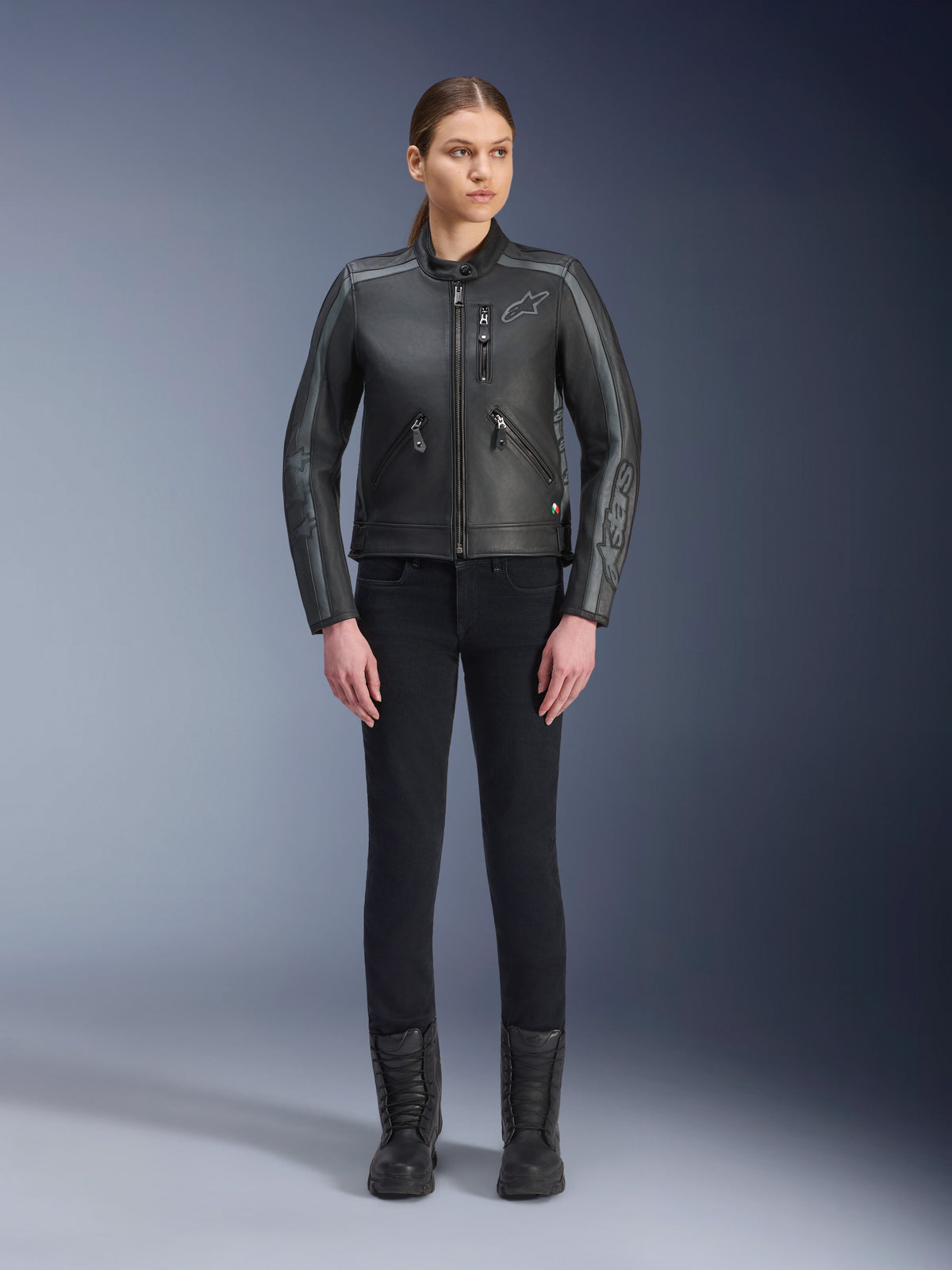 Alpinestars Stella Dyno Leren Jas, dames motorjas, zwart tar grijs donkergrijs, studio-opname van het volledige lichaam van een model dat de jas draagt met een zwarte slim-fit broek en zwarte laarzen, vervaardigd uit runderleer met grijze accenten en subtiele Alpinestars-branding op de borst en mouwen.