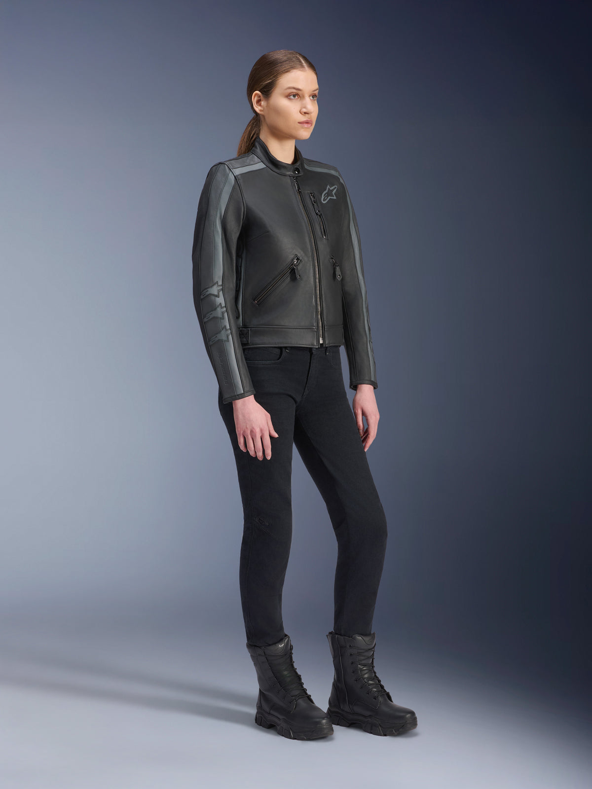 Alpinestars Stella Dyno Leren Jas, motorjas voor dames, Black Tar Gray en donkergrijs, gedragen met zwarte motorbroek en laarzen, voorzien van grijze racestrepen op de mouwen en erfgoed leerontwerp