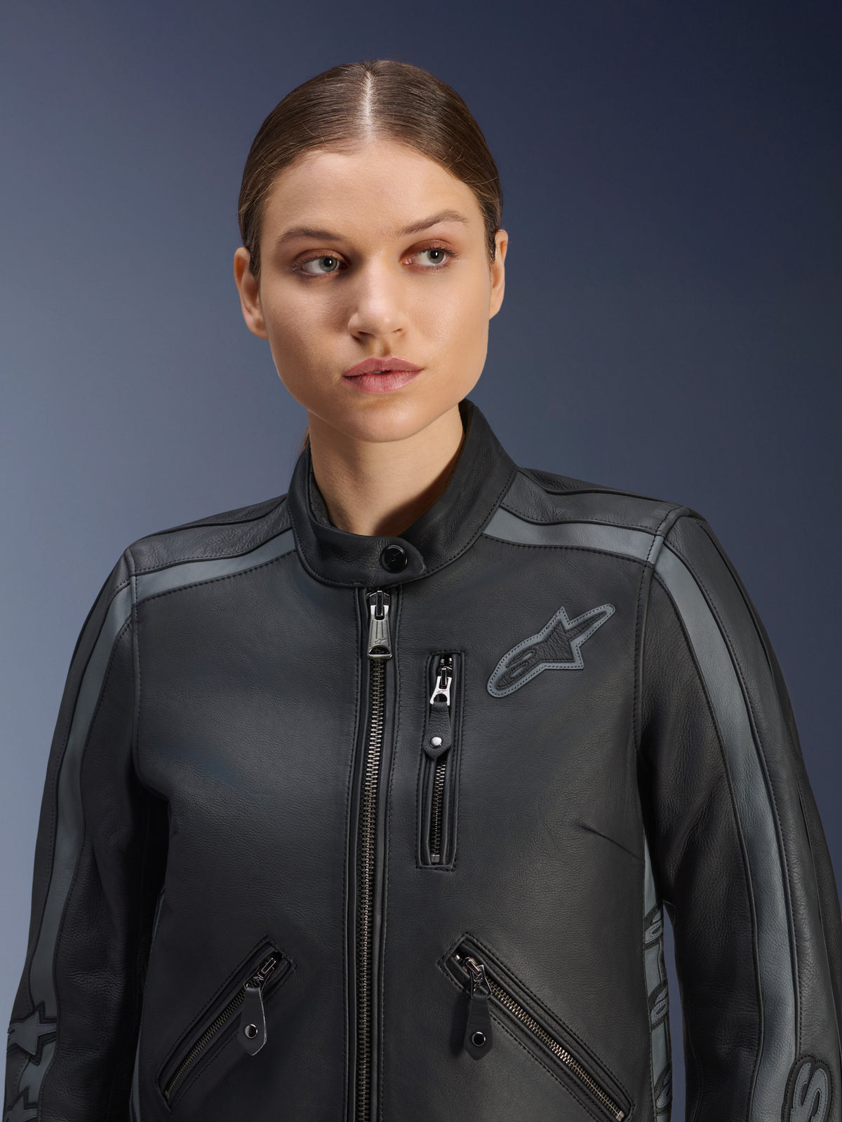 Vrouwelijk model draagt de Alpinestars Stella Dyno Leren Jas in Black Tar Gray Dark Gray, een premium runderleren motorjas met een erfgoedontwerp, voorzien van grijze schouderaccenten en meerdere zakken met ritssluiting, op maat gemaakt voor een specifieke pasvorm voor vrouwen.