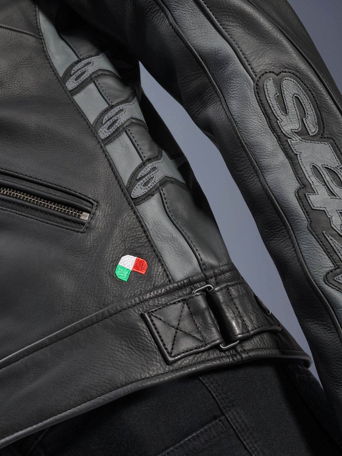 Alpinestars Stella Dyno leren jas detail, zwart en Tar Gray runderleer, close-up met klein geborduurde Italiaanse vlag, verstelbare gesp in de taille, grijze leren panelen met Alpinestars-logo's in reliëf en Stella-branding op de mouw