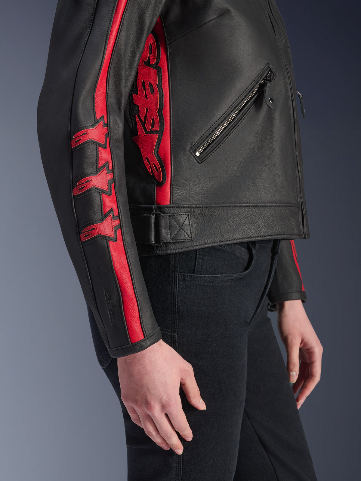 Alpinestars Stella Dyno leren jas, dames motorjas, zwart met Haute Red, zijaanzicht gedragen door een model, voorzien van rode racestrepen met sterlogo's op de mouw en een groot rood logo aan de zijkant, hoogwaardig runderleer, urban rijstijl