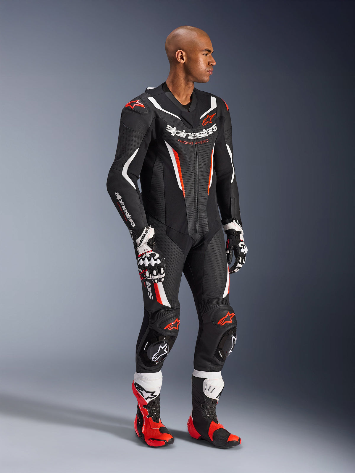 Modèle portant la combinaison en cuir Alpinestars GP Force V2 1 pièce en Noir Blanc Rouge Fluo, équipement de course haute performance avec panneaux en cuir perforé et boucliers DFS Lite, complétée par des bottes et des gants de course rouges et blancs assortis, esthétique sport mécanique professionnelle.