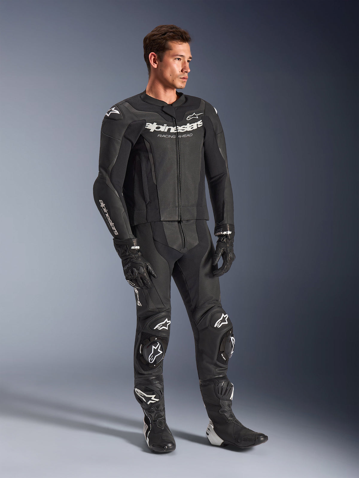 Combinaison en cuir 2 pièces Alpinestars GP Force V2, noire, portée par un mannequin homme avec gants et bottes de course noirs assortis, logos Alpinestars blancs, panneaux en cuir perforé pour la ventilation et sliders de genoux intégrés, équipement professionnel de course moto