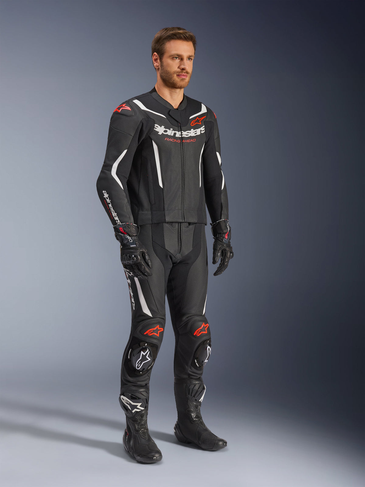 Alpinestars GP Force V2 2-teiliger Lederkombi, Schwarz, Weiß und Rot Fluo, getragen von einem Modell mit schwarzen Rennstiefeln und Handschuhen, mit perforierten Ledereinsätzen, weißen aerodynamischen Akzenten und roten Fluo-Logos an Schultern und Knien, professionelle Motorrad-Rennausrüstung