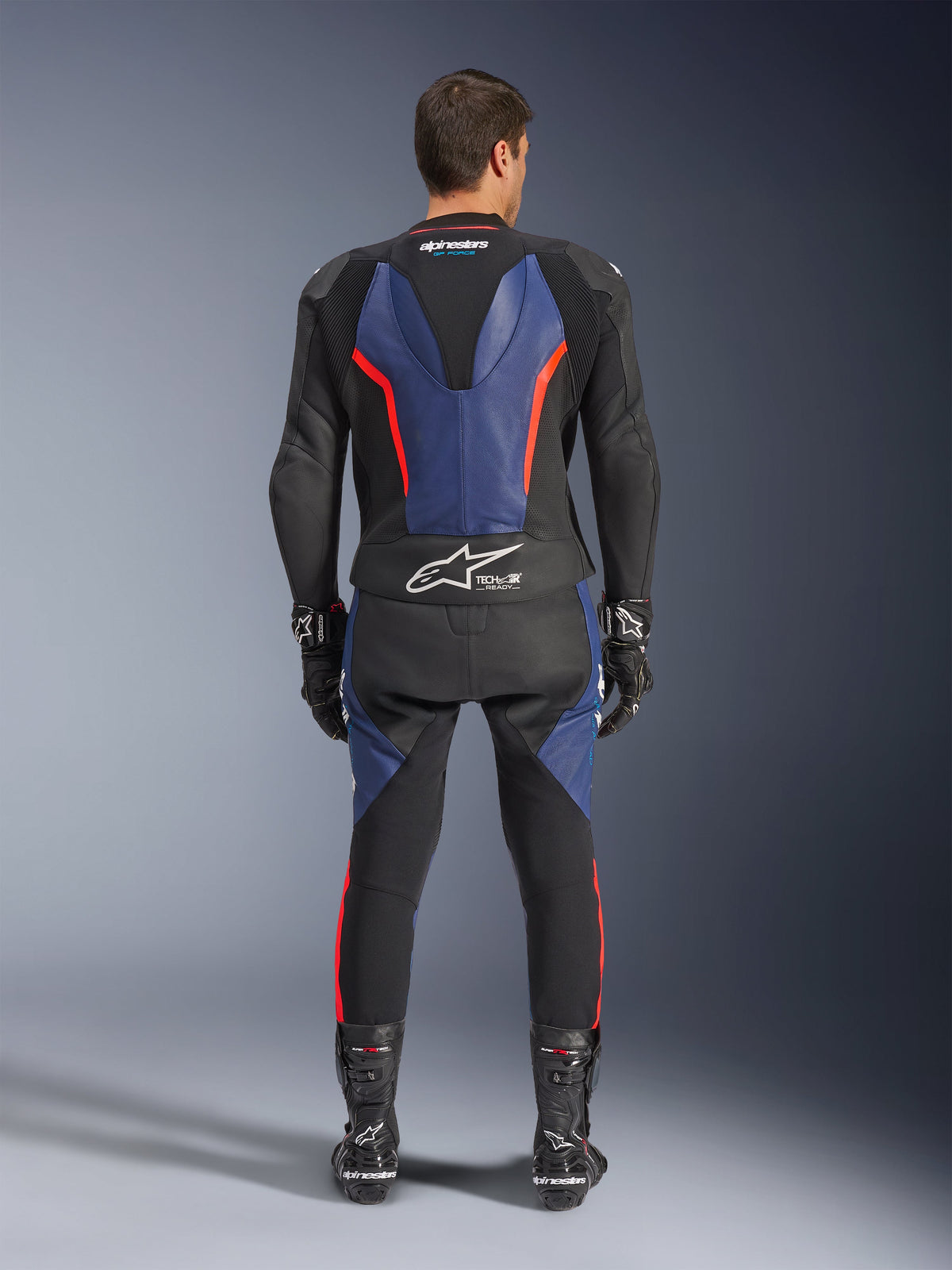 Alpinestars GP Force V2 Lederkombi 2-teilig, Rückansicht, Schwarz, Blau und Rot Fluo, mit blauen Lederbahnen an Rücken und Beinen sowie fluoreszierend roten Akzenten, Tech-Air Ready-Branding am unteren Rücken, getragen mit schwarzen Rennhandschuhen und Stiefeln.