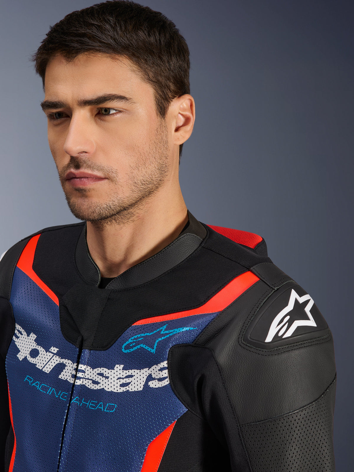 Alpinestars GP Force V2 Lederkombi – 2-teilig, Schwarz, Blau und Fluo-Rot, Nahaufnahme eines Models, das die Jacke trägt, mit perforierten Rindsledereinsätzen, Alpinestars-Logo auf der Schulter und „Racing Ahead“-Schriftzug auf der Brust, professionelle Rennausrüstung