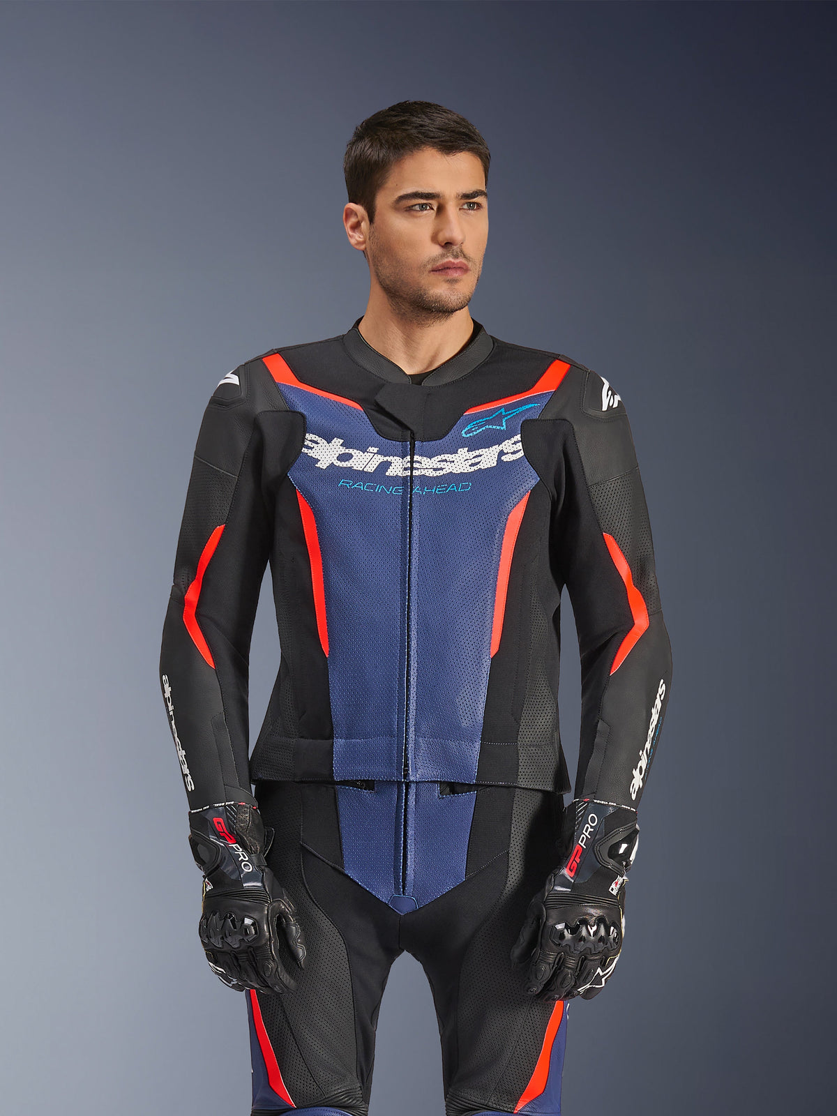 Männliches Model trägt Alpinestars GP Force V2 Lederkombi 2-teilig, Schwarz Blau Rot Fluo, mit markantem blauem Brustpanel und roten Akzenten, kombiniert mit GP Pro Handschuhen, professionelle Motorrad-Rennbekleidung aus Leder mit perforierter Textur