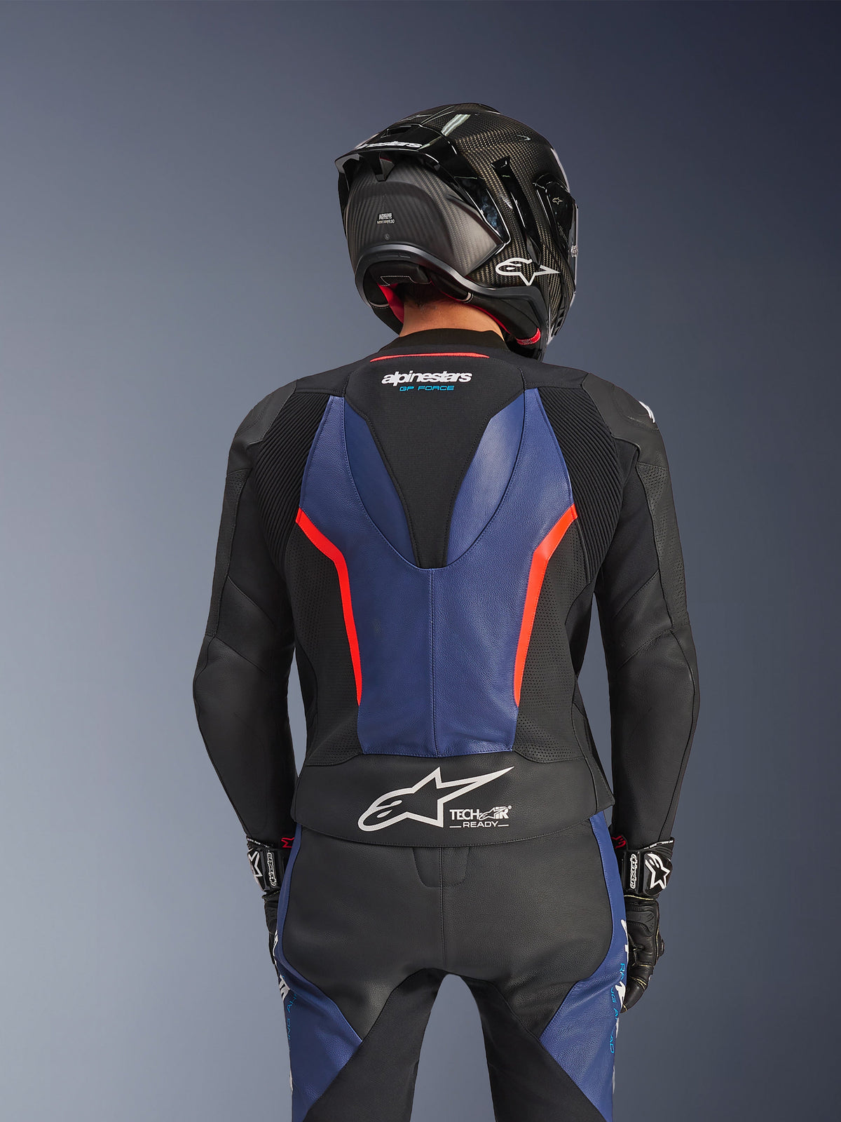 Alpinestars GP Force V2 2-teilige Lederkombi, Schwarz Blau Rot Fluo, Rückansicht mit aerodynamischem Speed-Hump und Tech-Air Ready Branding, hochwertiges Rindsleder mit Stretchzonen, kombiniert mit einem schwarzen Carbon-Helm, professionelle Motorrad-Rennausrüstung
