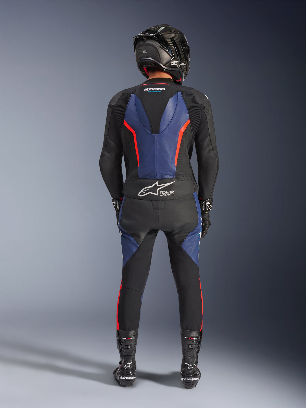 Alpinestars GP Force V2 Lederkombi 2-teilig, Schwarz Blau Rot Fluo, vollständige Rückansicht eines Fahrers in kompletter Rennausrüstung, mit blauen Lederbahnen am Rücken und an den Beinen sowie fluoreszierenden roten Akzenten, Tech-Air® Ready-Logo am unteren Rücken, kombiniert mit einem Carbon-Helm, schwarzen Handschuhen und Rennstiefeln.