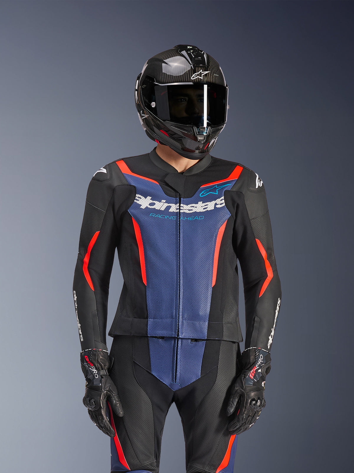 Alpinestars GP Force V2 Lederkombi 2-teilig, Schwarz, Blau und Rot Fluo, getragen mit einem Carbon-Helm und Rennhandschuhen, mit perforierten Ledereinsätzen und weißem Alpinestars-Logo auf der Brust, professionelle Renn- und Sportfahrausrüstung