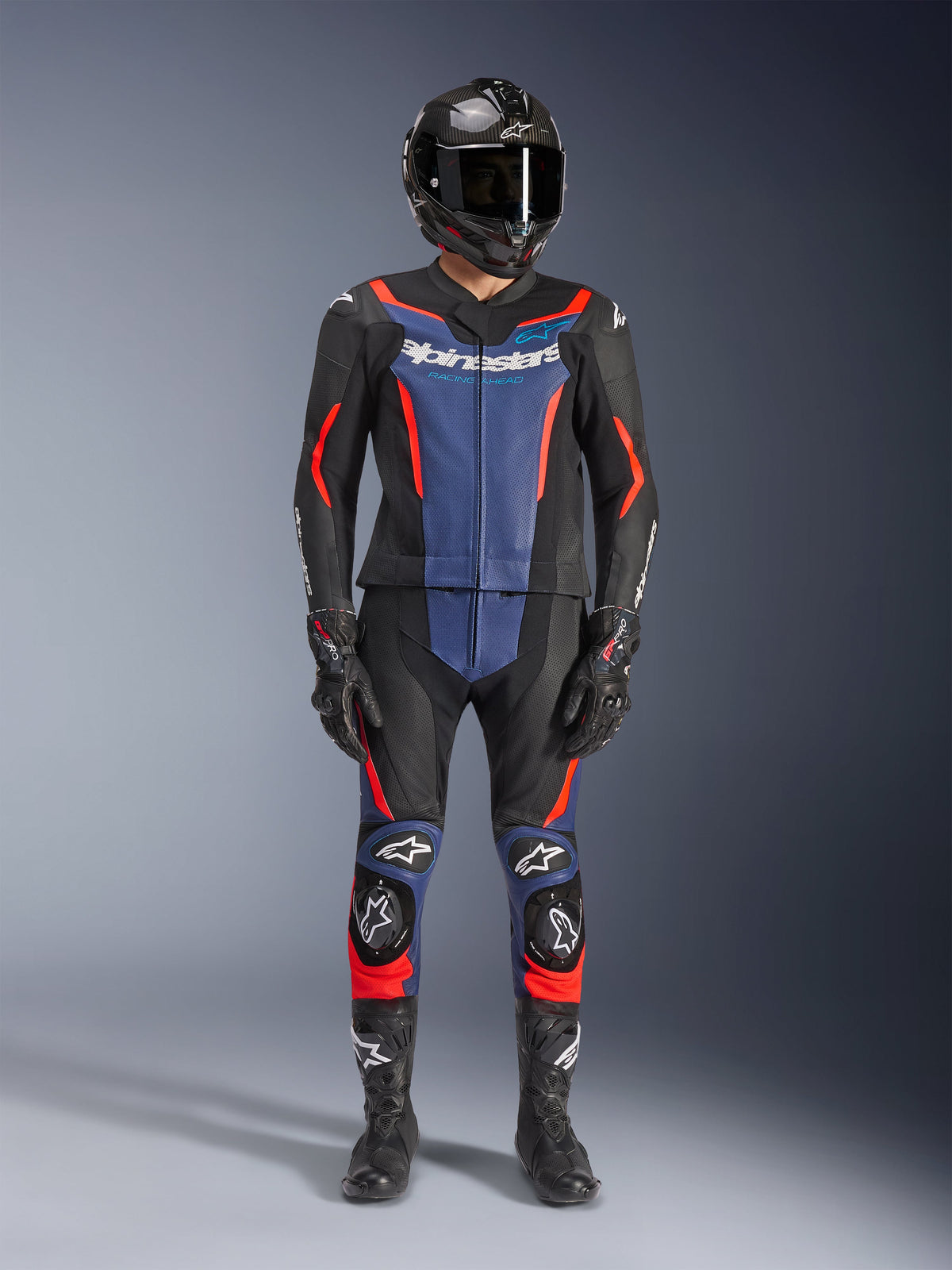 Alpinestars GP Force V2 Lederkombi – 2-teilig, Schwarz, Blau und Fluo-Rot, getragen mit einem Carbon-Integralhelm, schwarzen Rennhandschuhen und Stiefeln, mit blauen Brusteinsätzen und fluoreszierenden roten Akzenten, professionelle Motorrad-Rennausrüstung