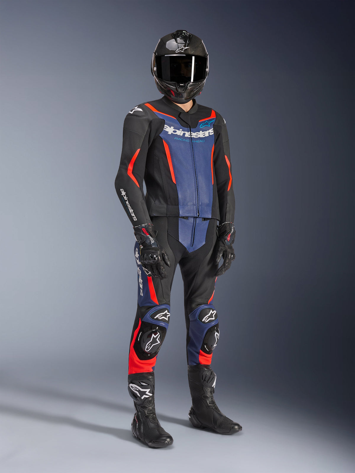 Alpinestars GP Force V2 Lederkombi – 2-teilig in Schwarz, Blau und Red Fluo, getragen mit einem Carbonfaser-Helm, schwarzen Rennhandschuhen und Stiefeln, mit perforierten Lederbahnen und weißem Alpinestars-Logo, professionelle Sport-Motorradausrüstung.
