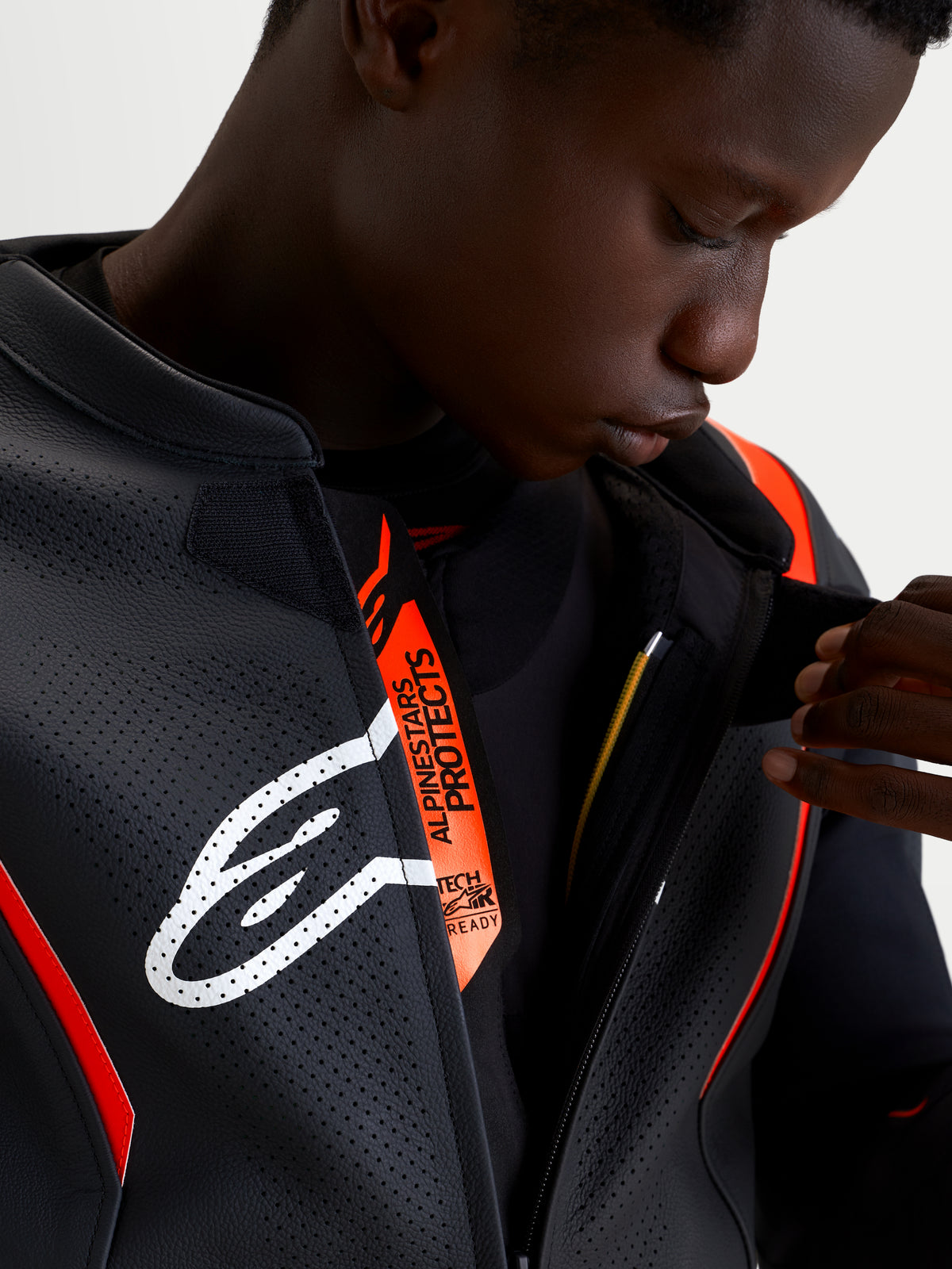 Alpinestars Missile V3 2 pièces, gros plan d'un modèle portant la combinaison en cuir noir et rouge fluo, montrant la patte de fermeture avant perforée avec le logo Astar blanc et la mention Alpinestars Protects