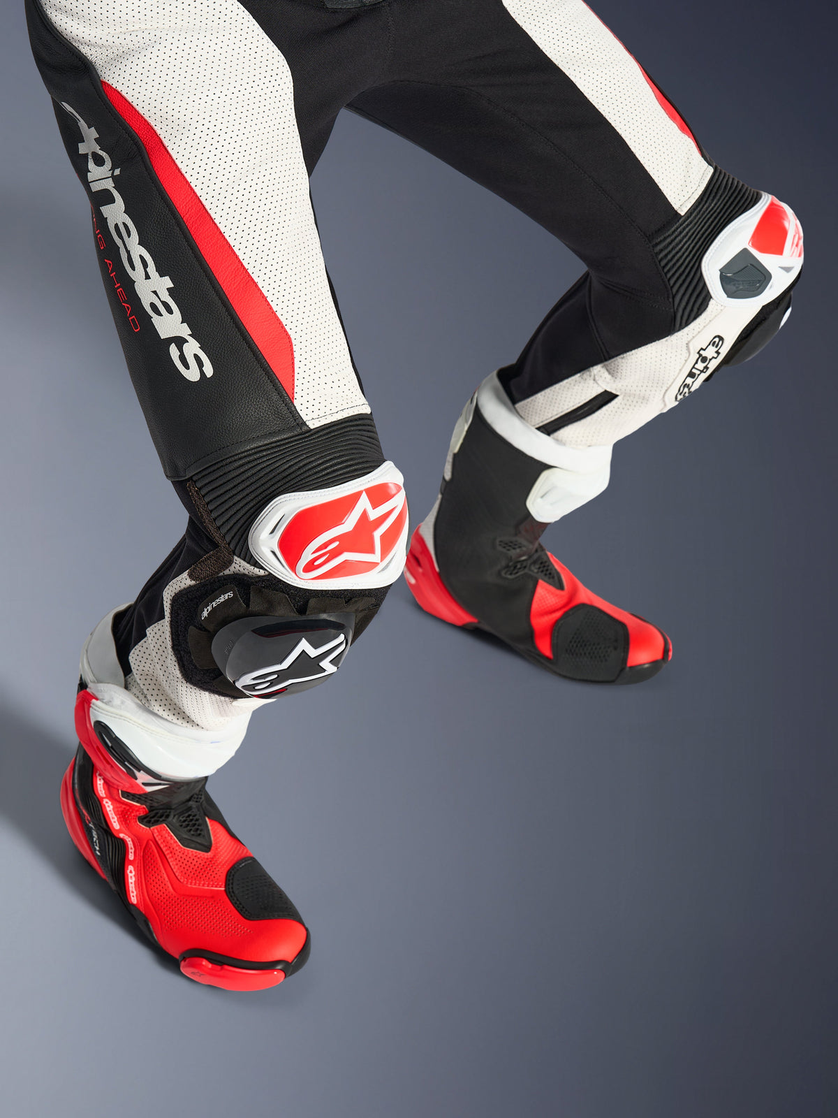 Alpinestars Stella GP-R7 2-delig dameslederen pak, onderbenen en racelaarzen, zwart, wit en fluo rood, voorzien van geperforeerd leer, opvallend Alpinestars-logo op het bovenbeen, accordeon-stretchpanelen en rood-witte kniesliders met bijpassende rood-zwarte technische laarzen.