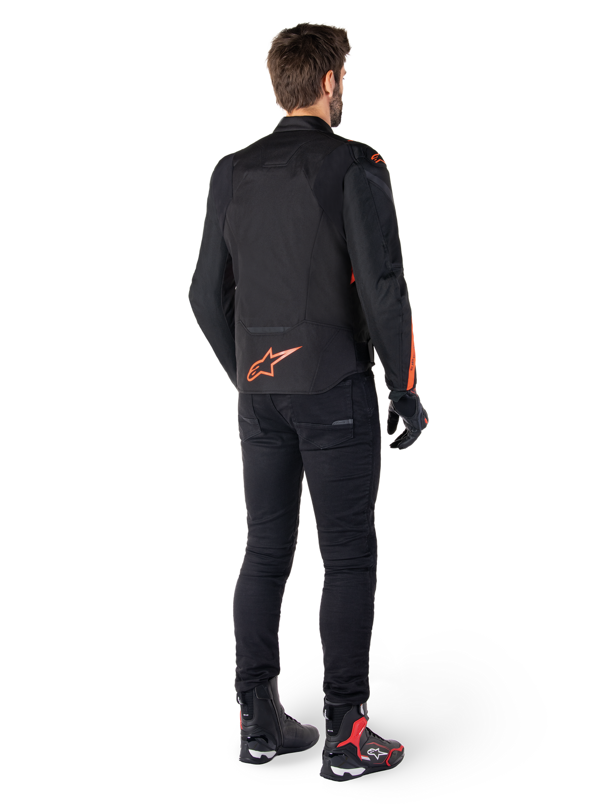Veste imperméable Alpinestars T-Jaws V4, noir et rouge fluo, vue de dos portée par un motard, présentant un logo rouge fluo éclatant dans le dos, associée à un pantalon de moto noir et des bottes techniques, équipement de moto sportif.