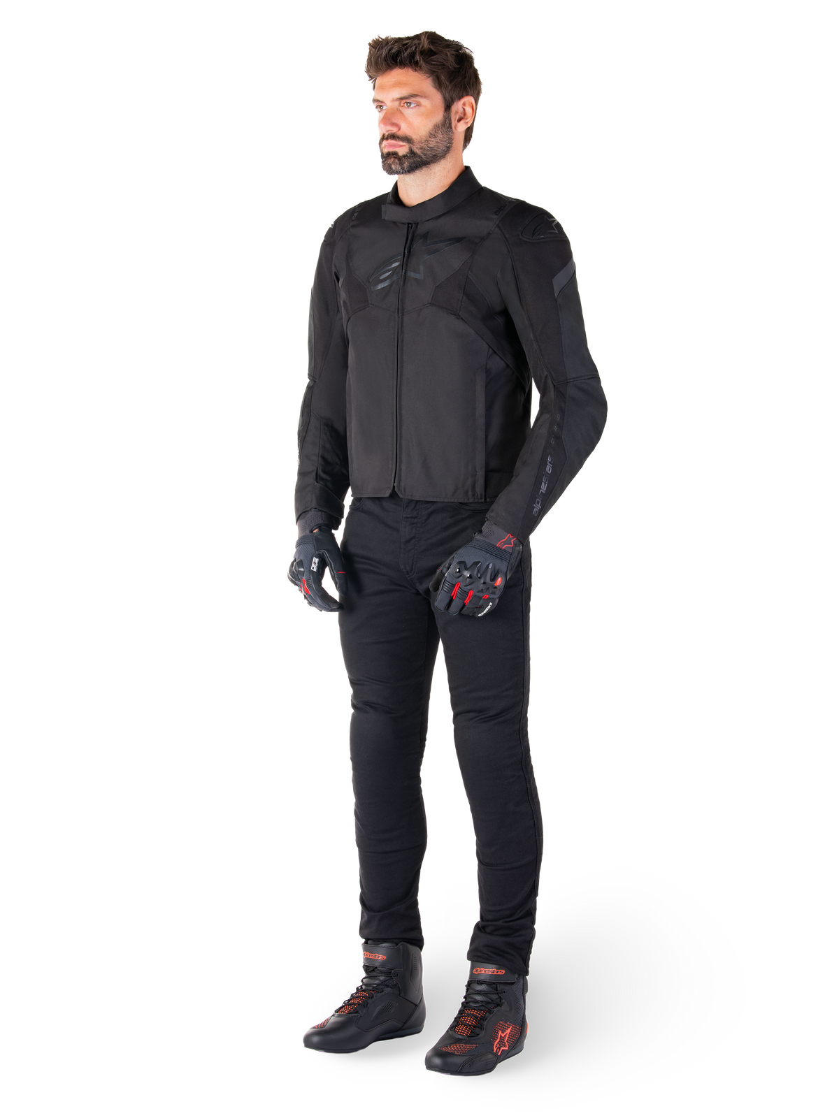 Männliches Modell in der Alpinestars T-Jaws V4 Wasserdichten Jacke in Schwarz, kombiniert mit schwarzer Motorradhose, schwarzen Handschuhen mit roten Akzenten und schwarzen Motorradschuhen, sportliche Passform, dezentes Branding, weißer Hintergrund