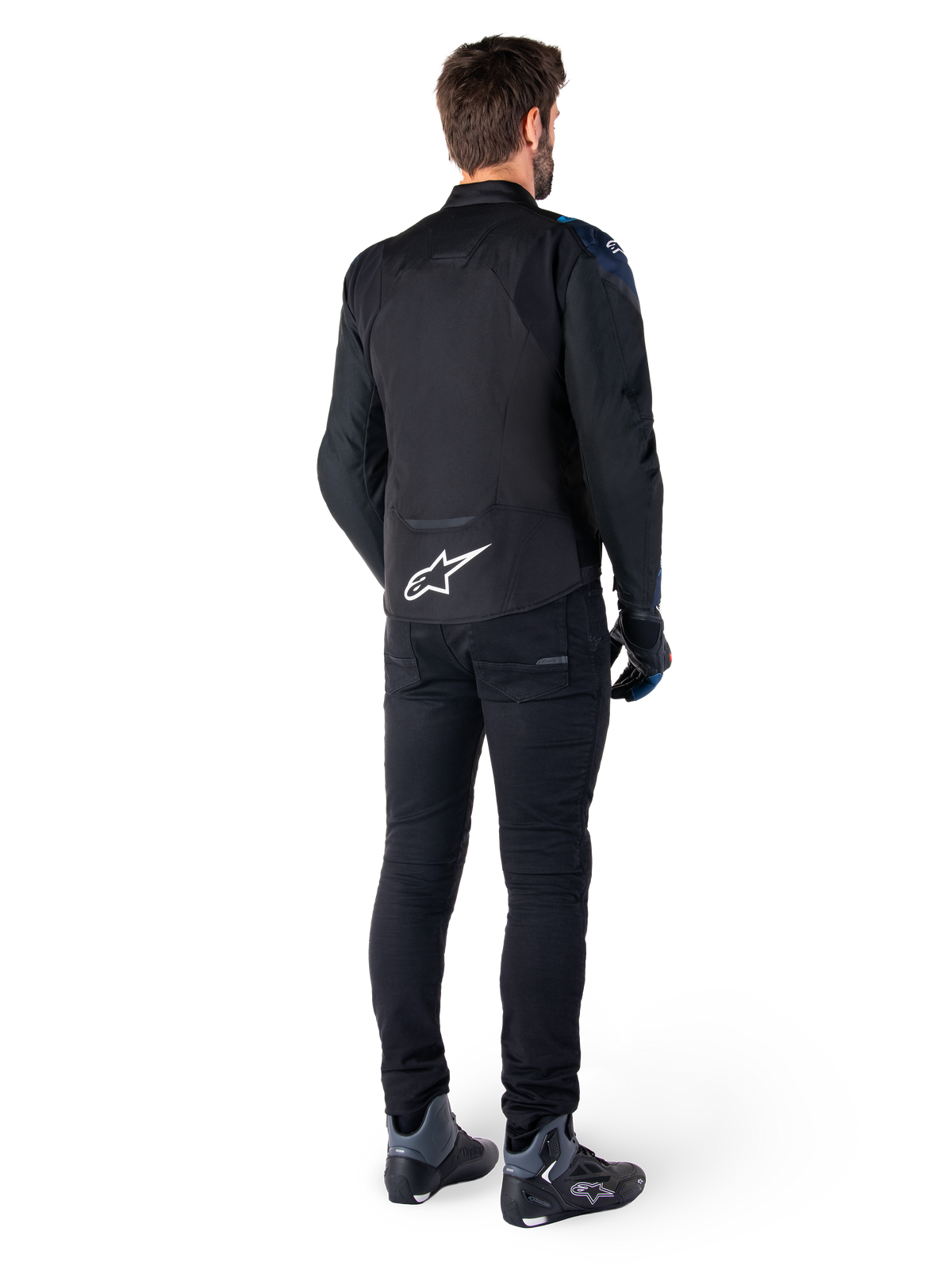 Vue arrière d'un homme portant la veste imperméable Alpinestars T-Jaws V4 en Noir, Bleu Foncé et Bleu Blithe, arborant un logo Astar blanc distinctif sur le bas du dos, associée à un pantalon noir et des chaussures de moto.