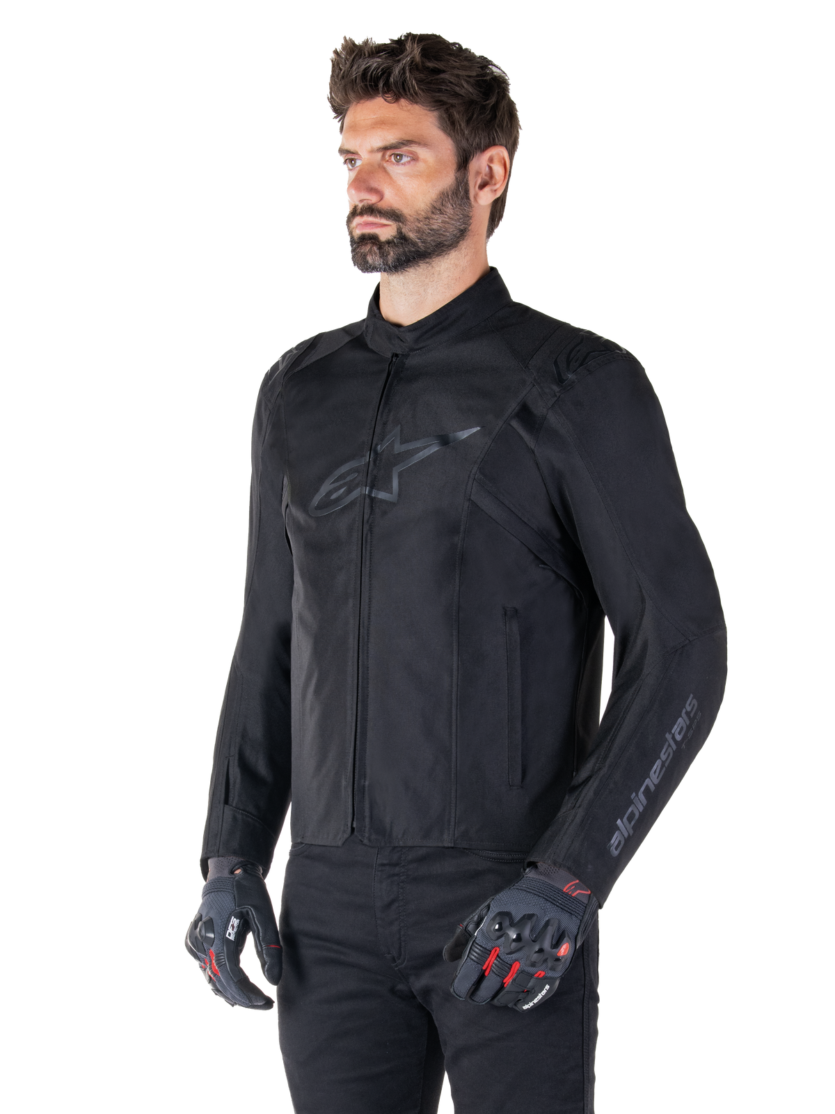 Model wearing Alpinestars T-SPS V2 Waterproof Jacket, Black, paired with black pants and grey motorcycle gloves with red accents, featuring a subtle black chest logo and sleeve branding, textile sport-cut jacket for road riding.