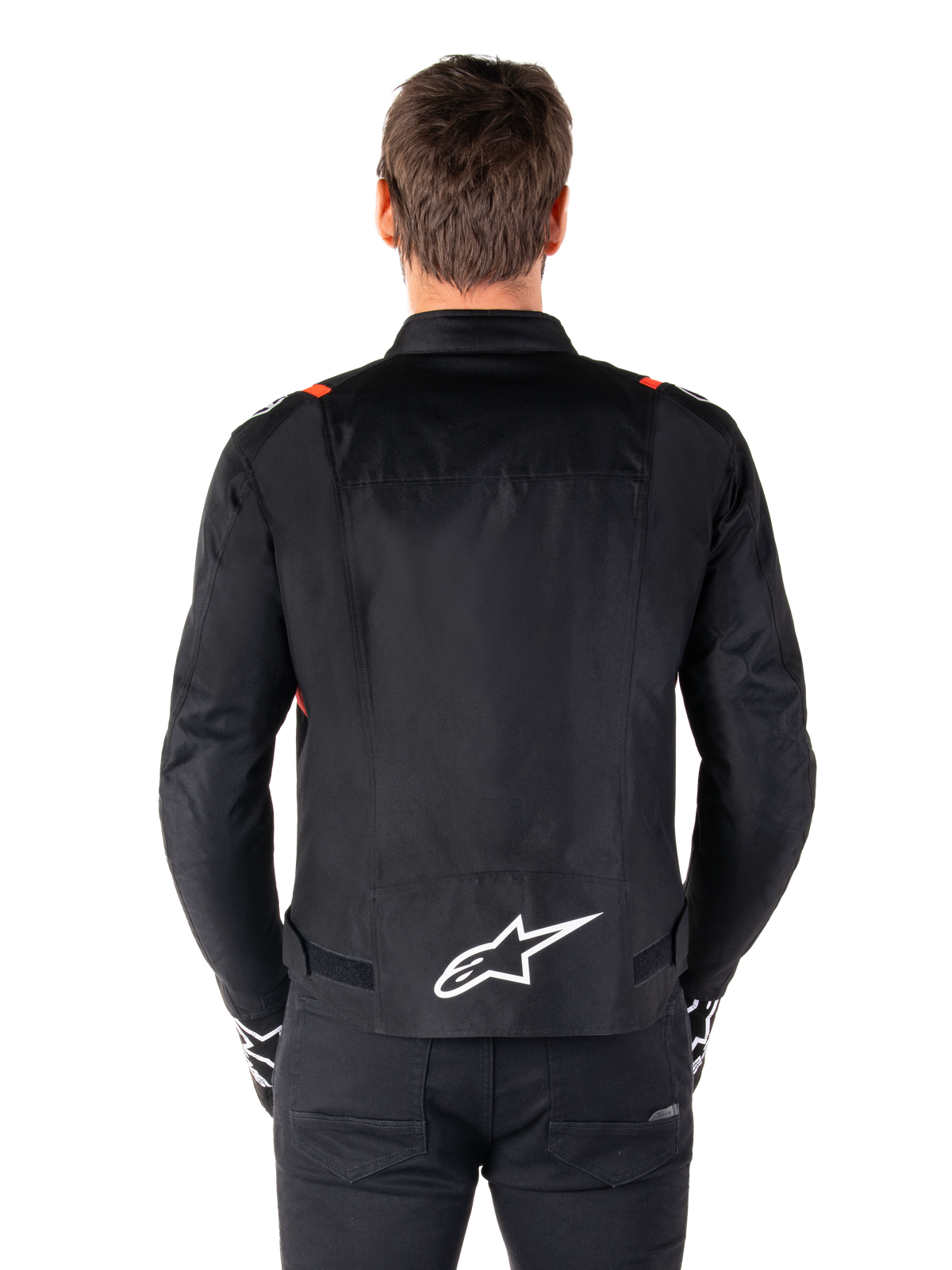 Alpinestars T-SPS V2 Wasserdichte Jacke, Schwarz mit weißen und fluoreszierend roten Akzenten, Rückansicht mit einem großen weißen Alpinestars-Logo am unteren Rücken, Textil-Motorradjacke im Sportschnitt, getragen mit schwarzer Hose und Handschuhen
