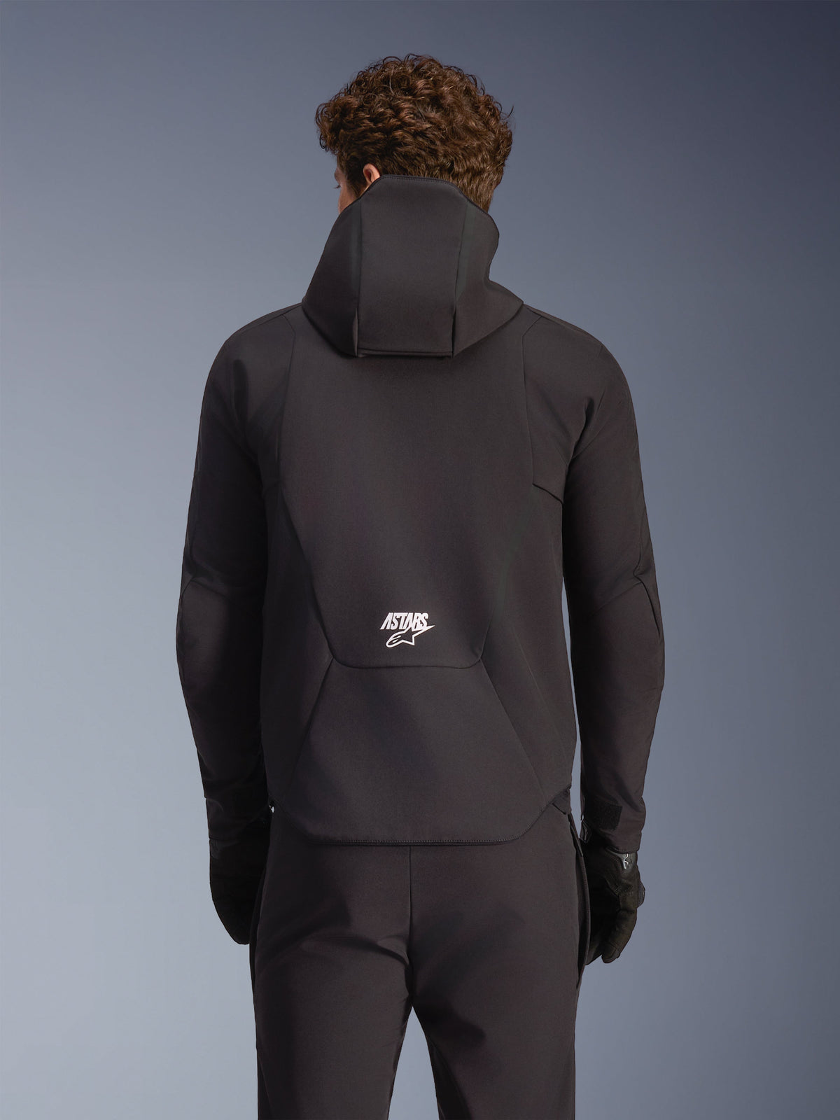 Alpinestars Aeroshell waterdichte jas, zwarte softshell motorjas, achteraanzicht met capuchon en wit logo op de onderrug.