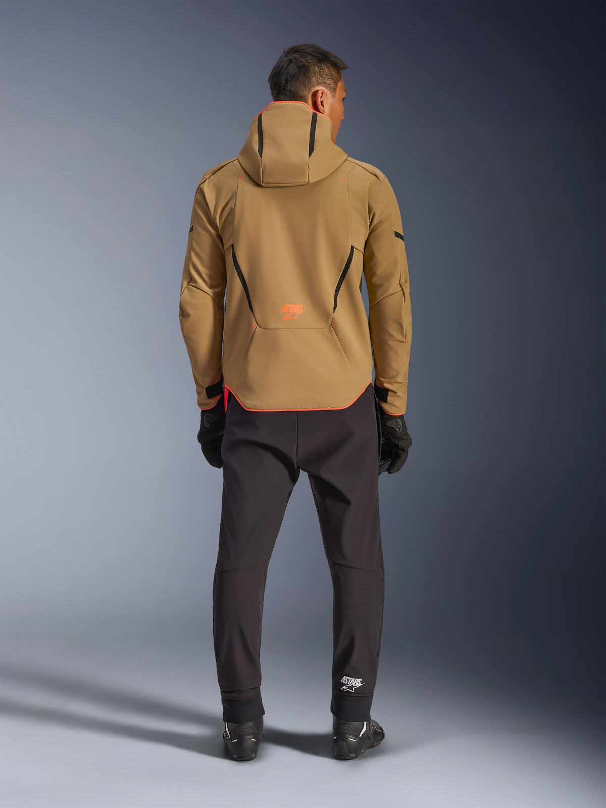 Alpinestars Aeroshell Waterproof Jacket in Dark Khaki Black Orange Fluo, achteraanzicht van een man die de softshell-jas met capuchon draagt, voorzien van zwarte biesjes en een oranje logo, gecombineerd met een zwarte technische broek en handschoenen.