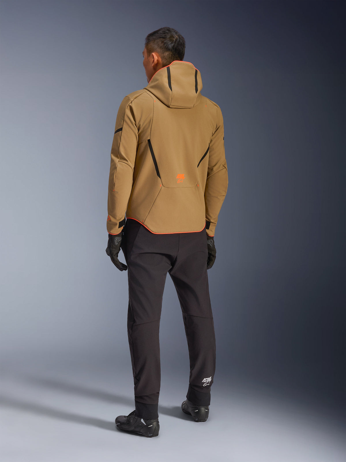 Alpinestars Aeroshell Waterdichte Jas, Dark Khaki Black Orange Fluo, achteraanzicht van een motorrijder die de softshell-jas met capuchon draagt, gecombineerd met een zwarte broek en handschoenen, voorzien van zwarte geometrische accenten en een oranje logo op de onderrug.