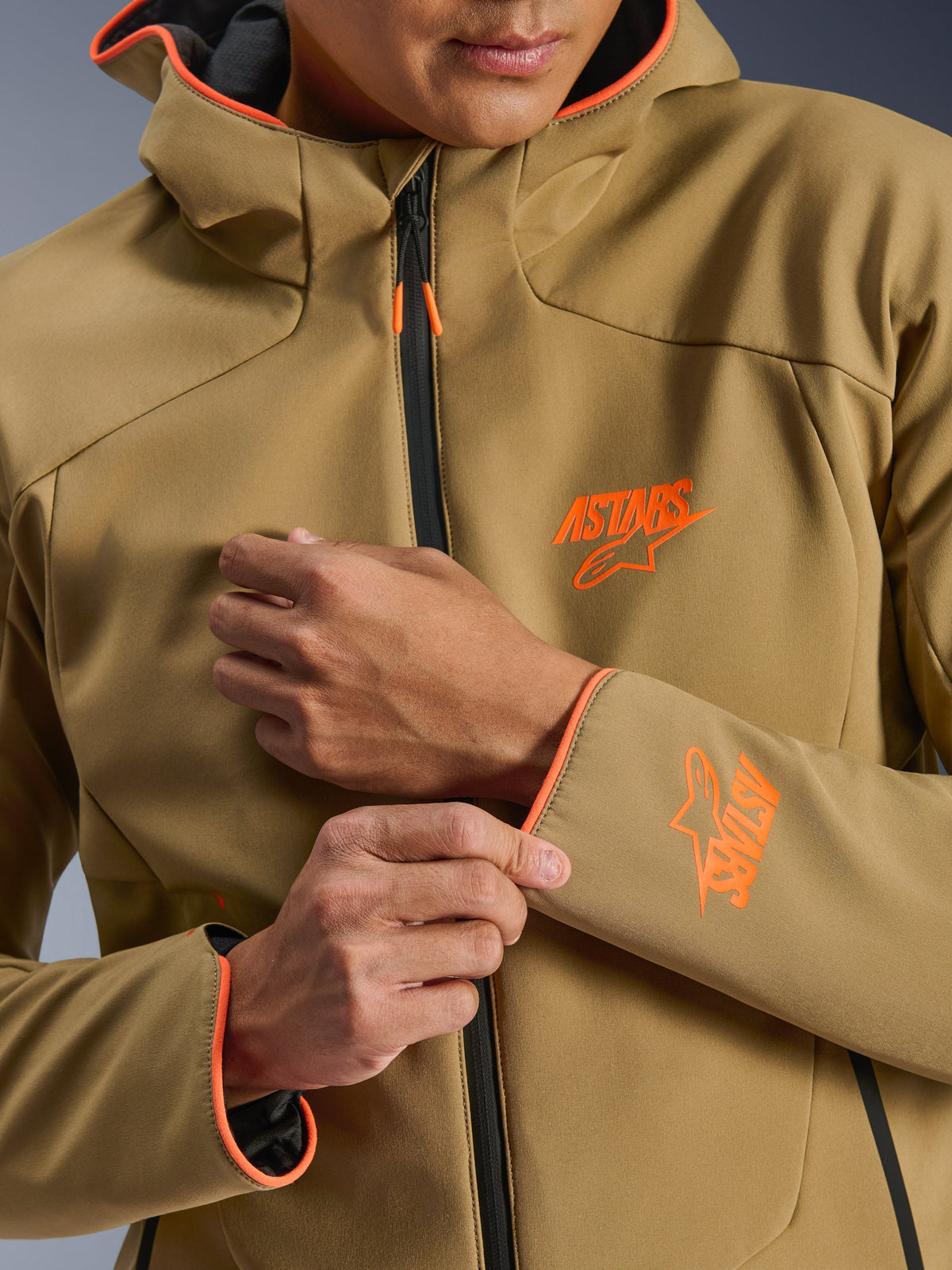 Alpinestars Aeroshell Waterproof Jacket, Dark Khaki en Orange Fluo, close-up van de voorzijde en mouw met oranje ASTARS-logo's, oranje biezen op de capuchon en de manchetten, en een zwarte waterdichte rits.