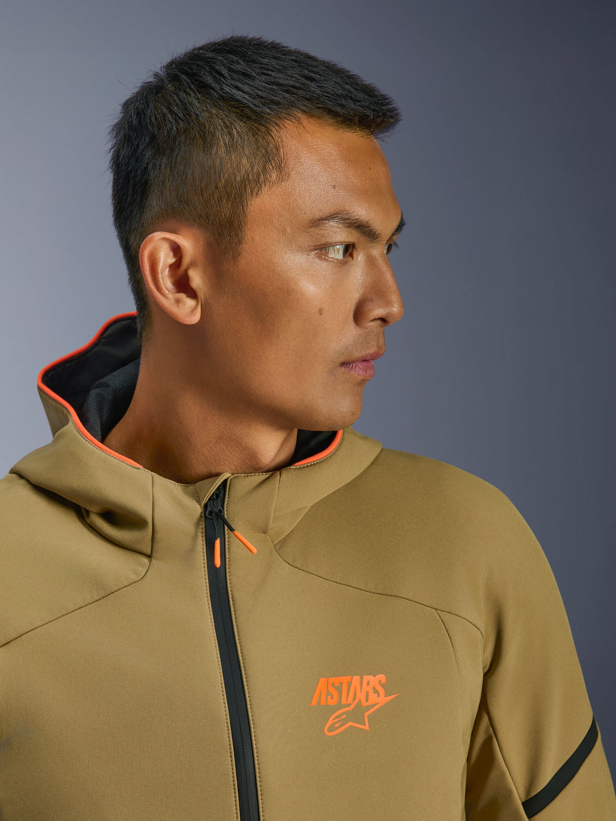 Alpinestars Aeroshell Waterproof Jacket, Dark Khaki en Orange Fluo, close-up zijprofiel, oranje ASTARS-logo op de borst en oranje gevoerde capuchon, moderne crossover softshell hoodie voor motorrijders