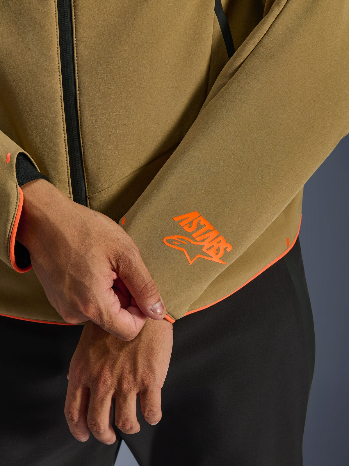 Alpinestars Aeroshell Waterproof Jacket, donker kaki met fluoriserend oranje accenten, close-up van de mouwboord en onderste zoom met het feloranje Astars-logo, neon oranje piping en detail van de waterdichte rits, sportieve softshell-constructie.