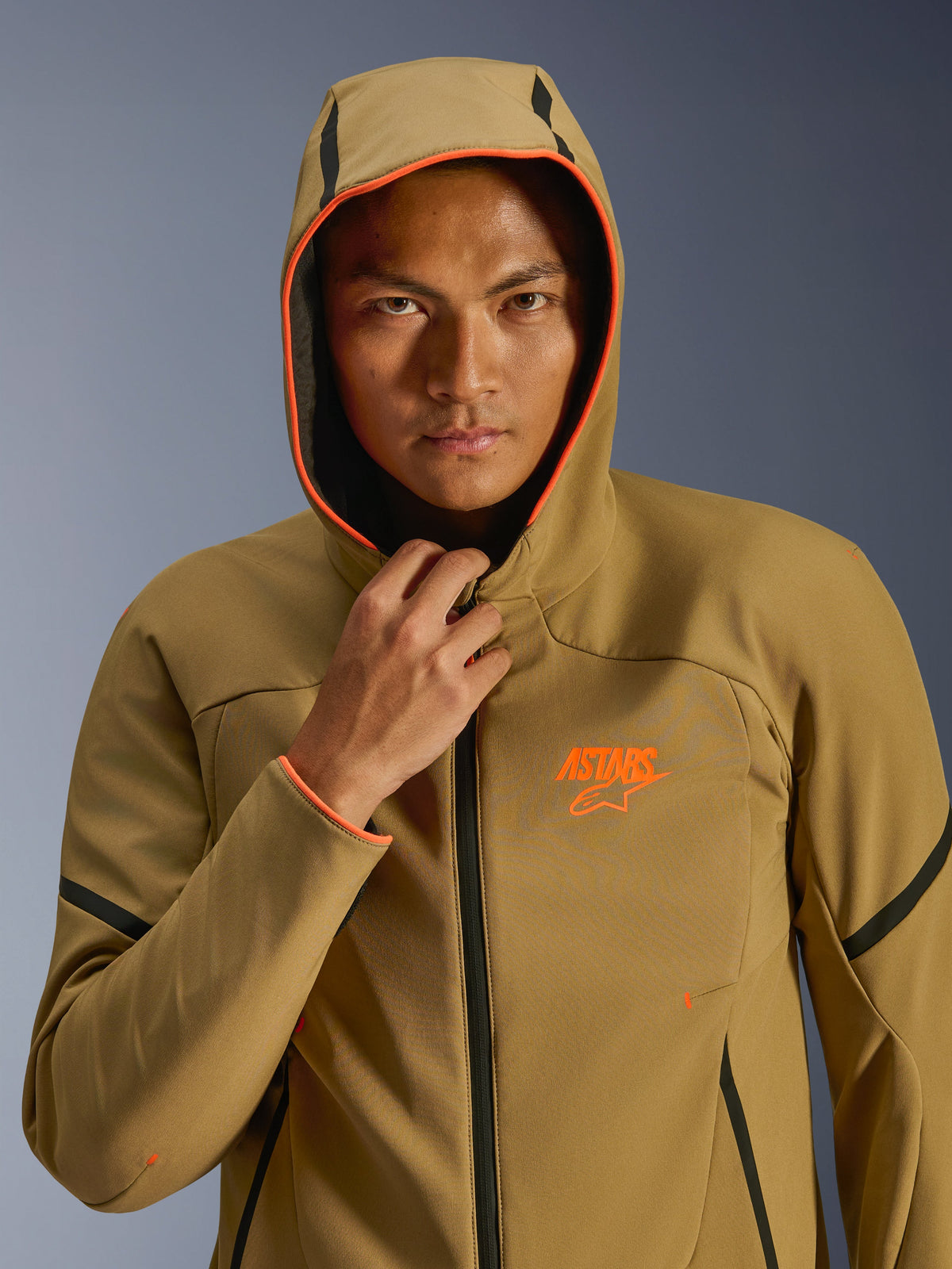 Alpinestars Aeroshell waterdichte jas, Dark Khaki Black Orange Fluo, model draagt de jas met de capuchon omhoog, voorzien van opvallende oranje fluo details en een oranje ASTARS-logo, strakke zwarte accenten, moderne sportieve waterdichte softshell