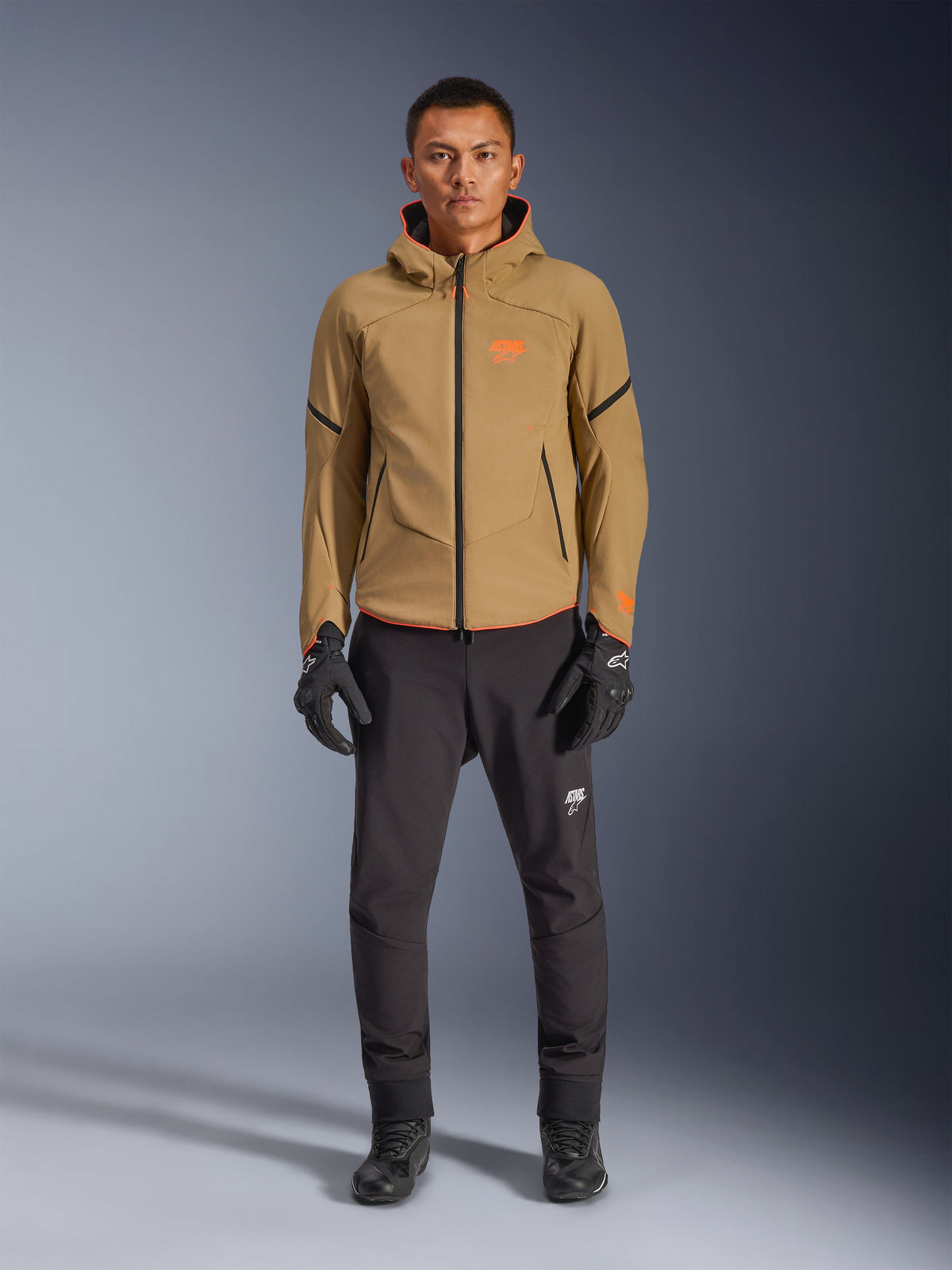 Mannelijk model draagt Alpinestars Aeroshell Waterproof Jas in Dark Khaki met zwarte en oranje fluo-accenten, oranje logo op de borst en ontwerp met capuchon, gecombineerd met een zwarte broek en zwarte motorhandschoenen, casual sport-touring stijl