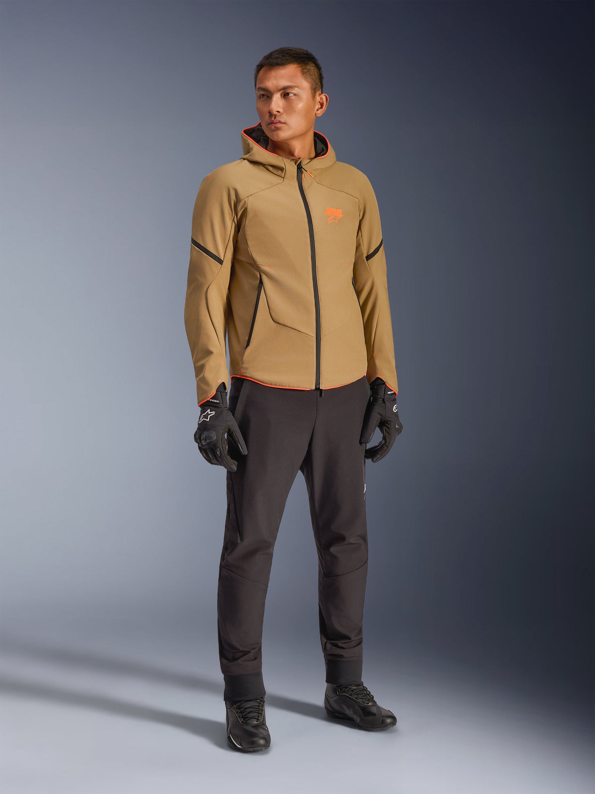Alpinestars Aeroshell Waterproof Jacket in Dark Khaki, zwart en oranje fluo, gedragen door een model met bijpassende uitrusting, softshell-ontwerp met capuchon, waterdichte ritsen en feloranje accenten en branding.