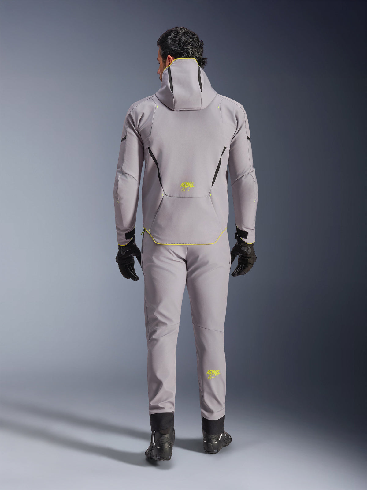 Alpinestars Aeroshell Waterproof Jacke in Mid Gray Yellow Fluo, vollständige Rückansicht, getragen mit passender grauer Hose, schwarzen Handschuhen und schwarzen Motorradschuhen. Verfügt über ein Kapuzen-Softshell-Design mit neongelben „ASTARS“-Logos und Akzenten, entwickelt für moderne Motorrad-Straßenperformance.